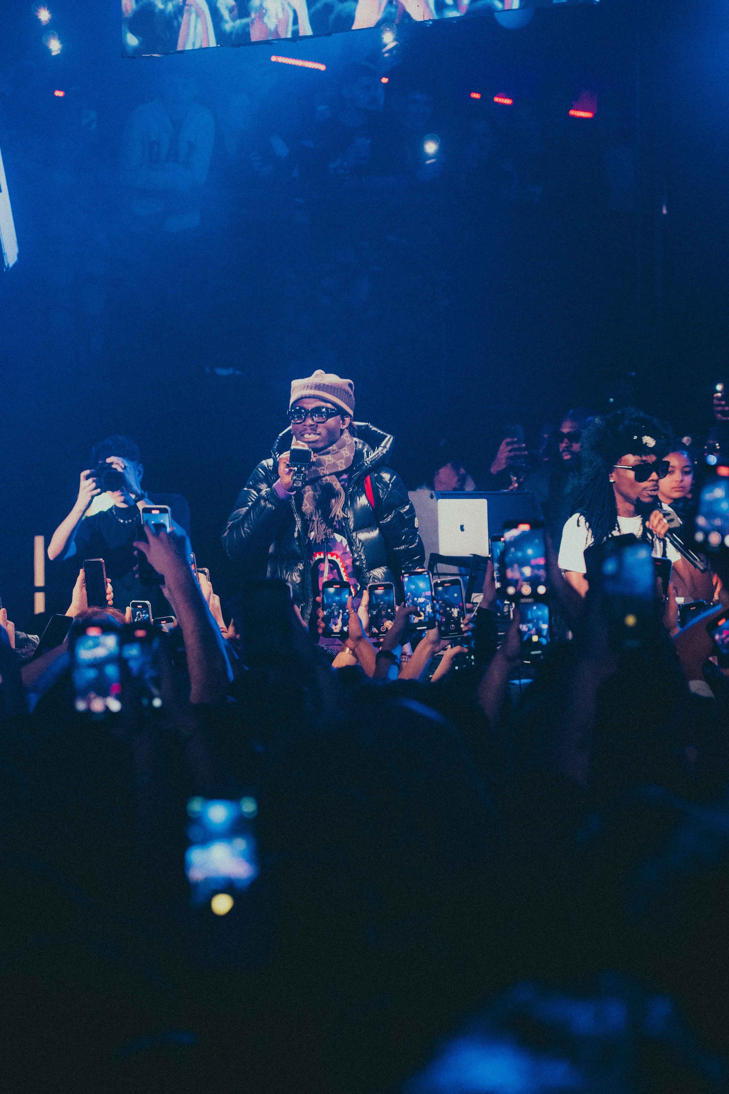 Un artiste ou un conférencier parle dans un micro lors d'un spectacle ou d'une événement, entouré de nombreux spectateurs qui prennent des photos avec leurs téléphones.