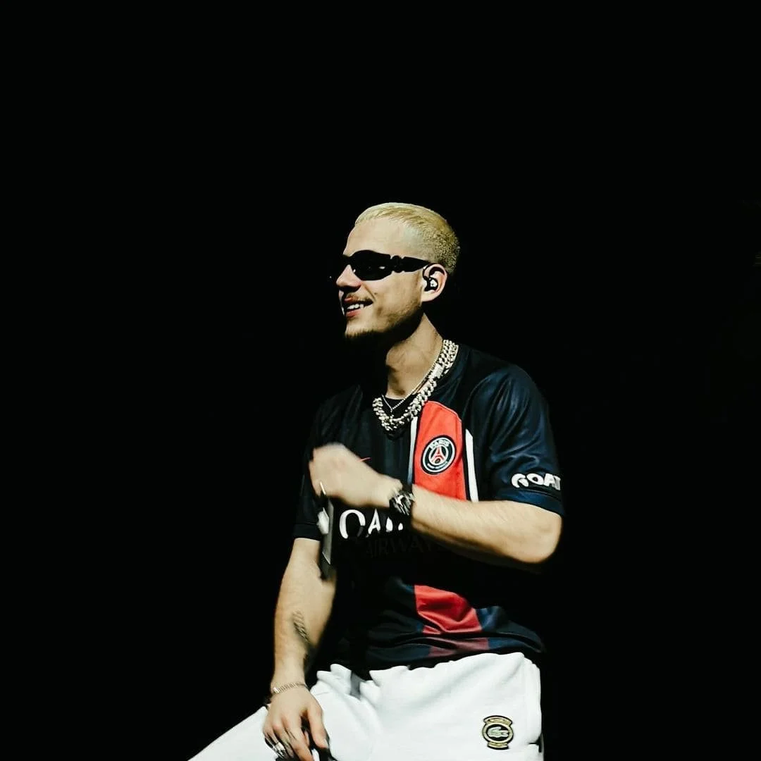 Un homme avec cheveux blonds platine portant des lunettes de soleil noires, habillé d'un maillot de football du Paris Saint-Germain, assis contre un fond noir, souriant et portant des bijoux
