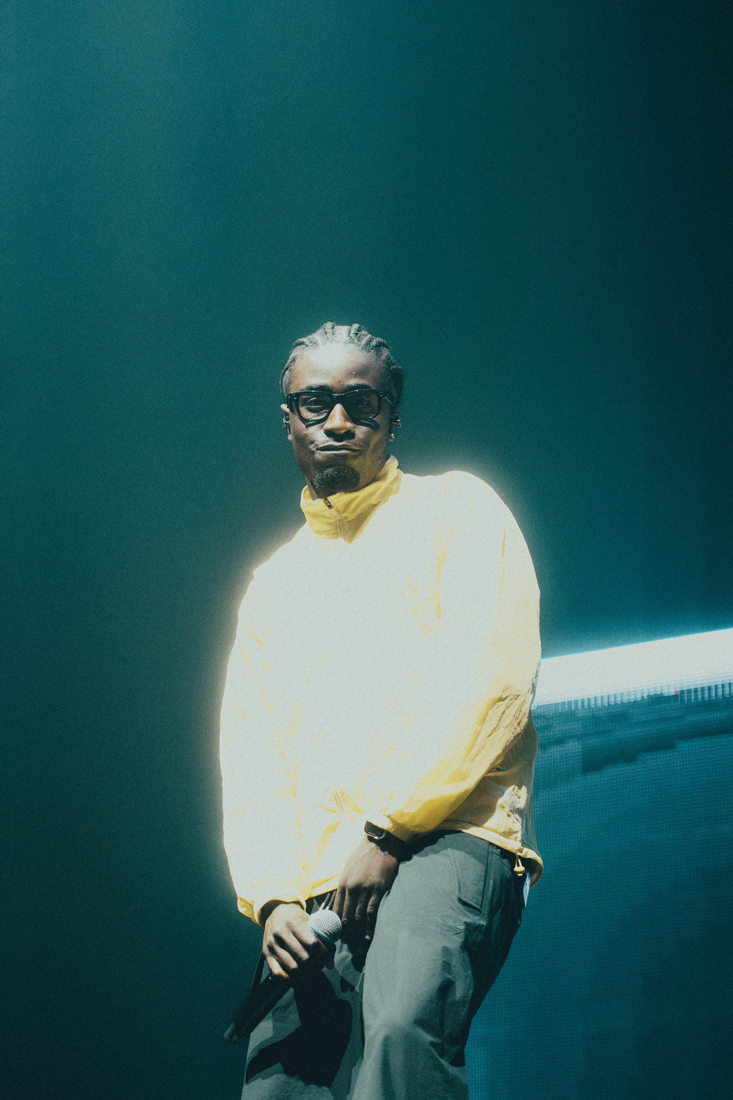 Jeune homme avec des dreadlocks portant des lunettes noires, un sweatshirt jaune, tenant un microphone, devant un fond lumineux et un écran LED.