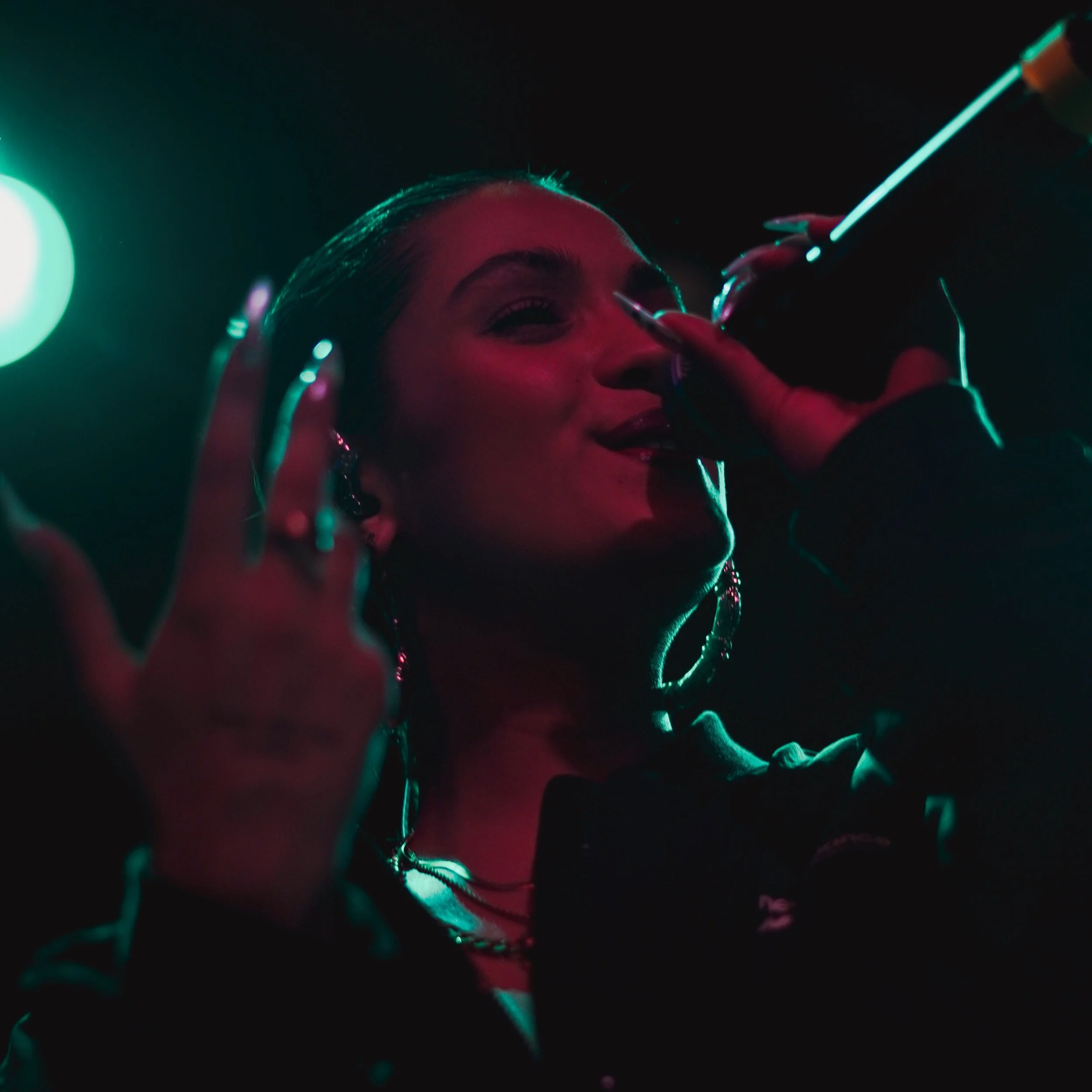 Une femme souriante avec de gros anneaux d'oreilles, recevant une boisson lors d'une fête ou d'un événement, sous un éclairage turquoise et rose.