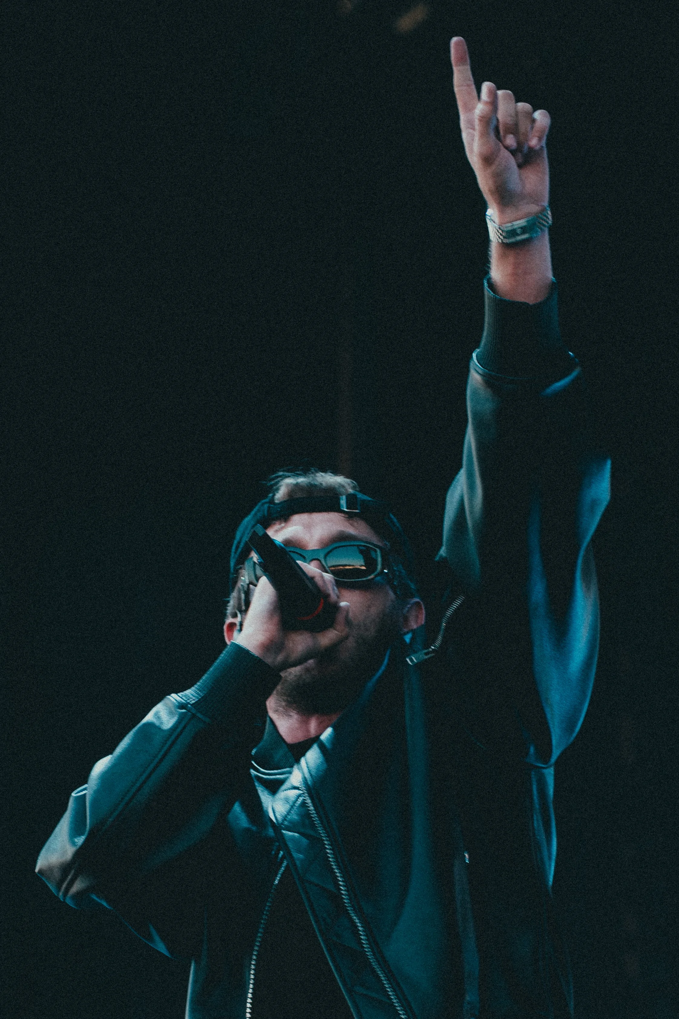 Un homme portant des lunettes de soleil, un casque et une veste en cuir, chante ou parle dans un microphone avec une main levée en l'air.