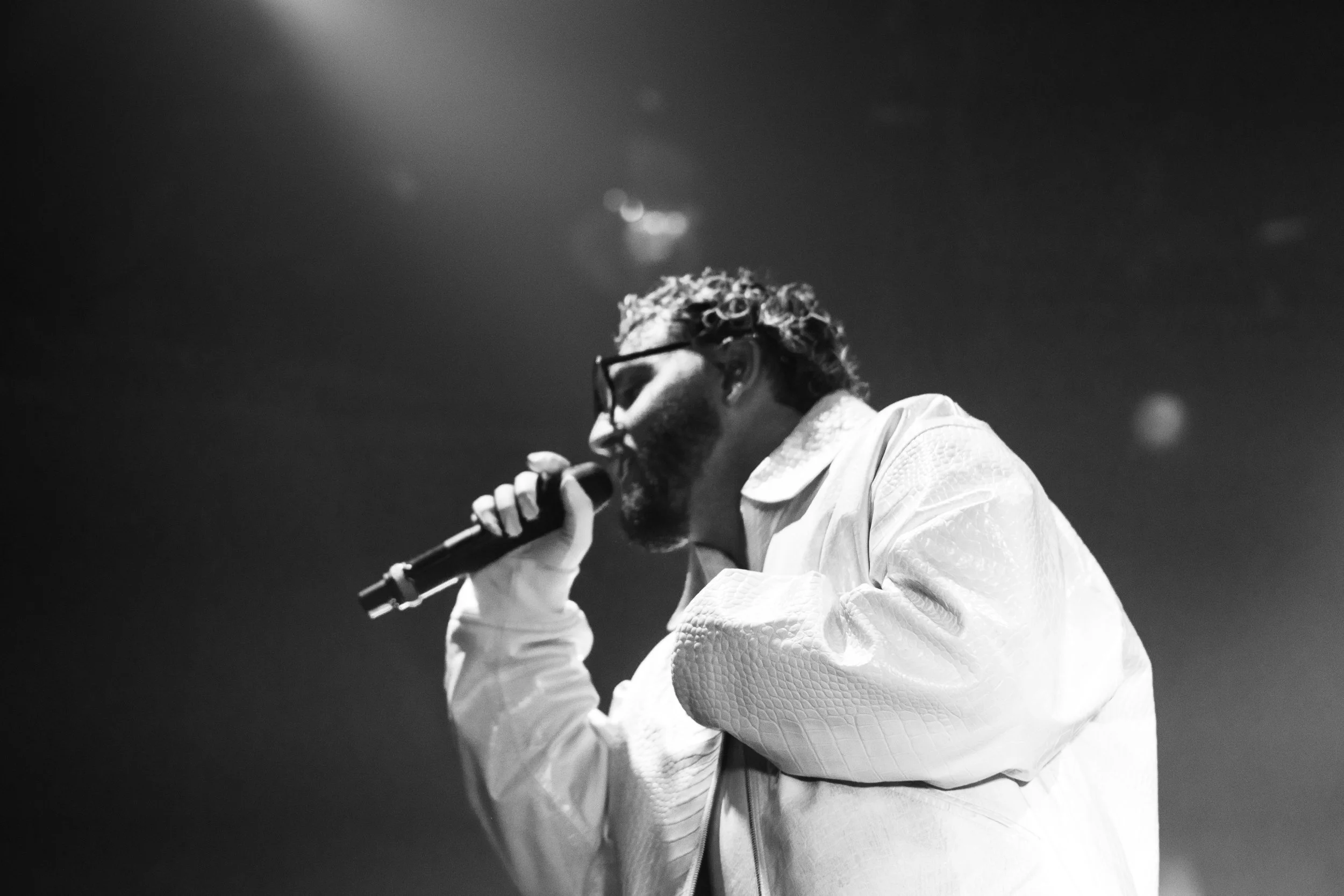 Un homme avec des lunettes, une barbe et des cheveux bouclés, portant une veste claire, chante dans un micro lors d'une performance sur scène, en noir et blanc.