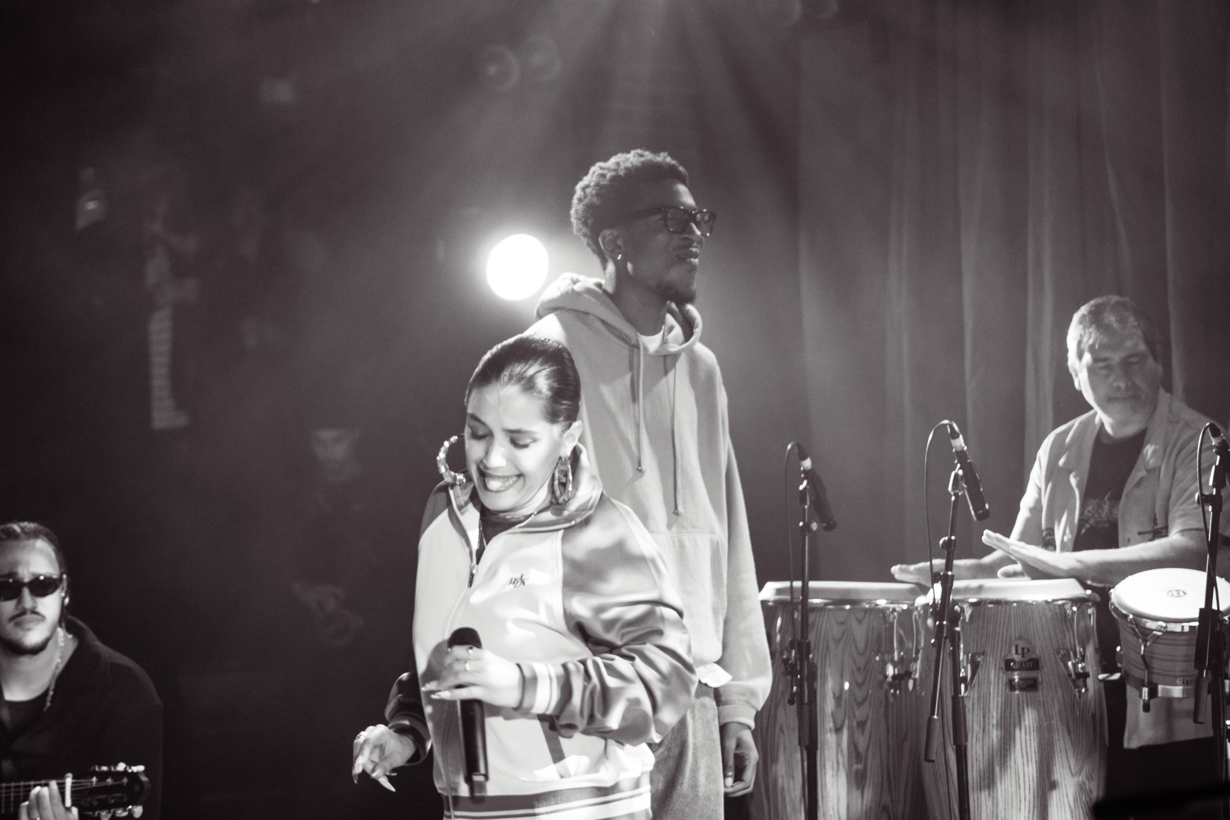 Un groupe de musiciens en performance sur scène, dont une femme chantant avec un microphone, un homme jouant de la percussion et un autre à la guitare, dans une ambiance sombre avec éclairage de scène.