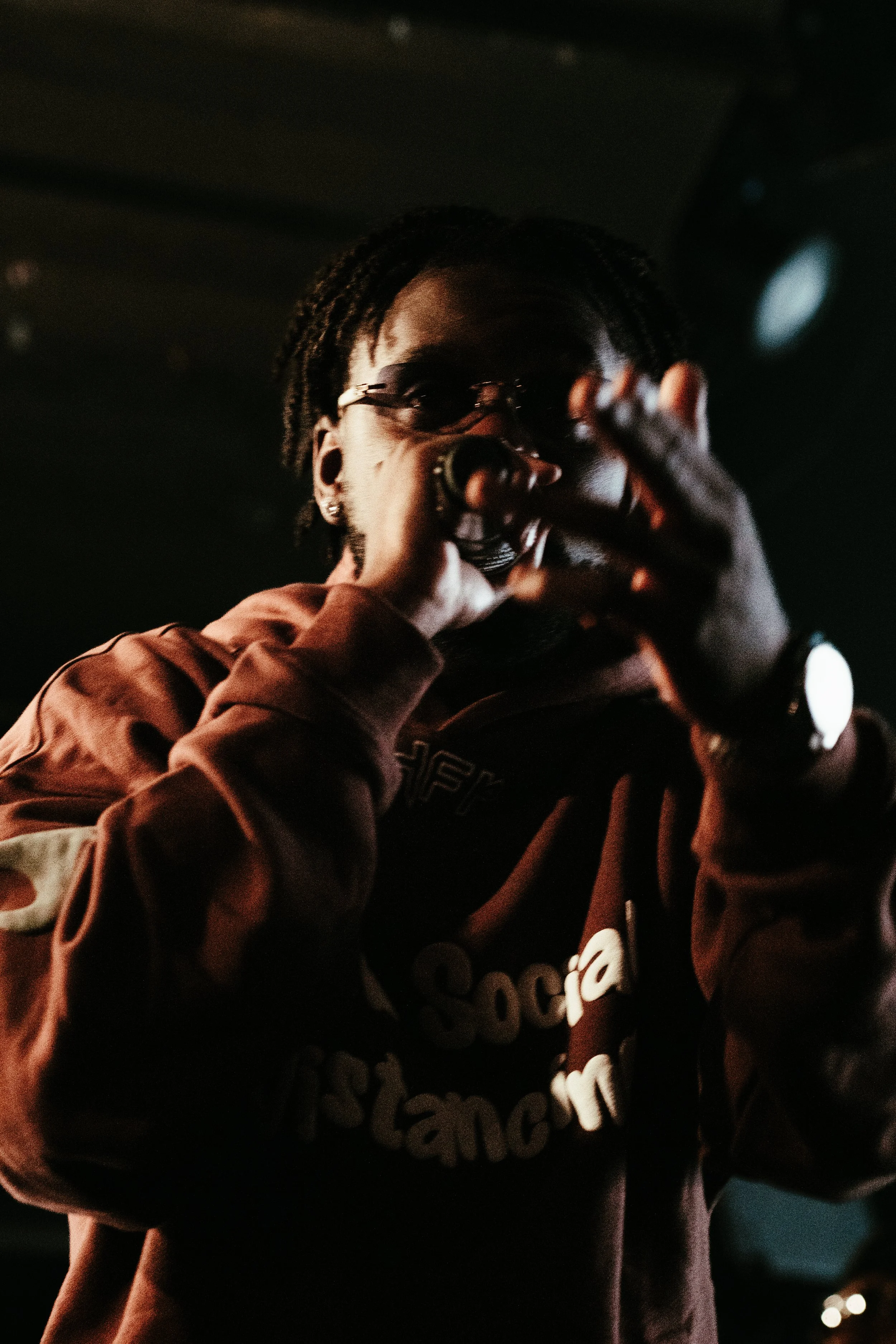 Jeune homme avec lunettes parlant ou chantant dans un endroit sombre, portant un sweat marron avec des inscriptions.