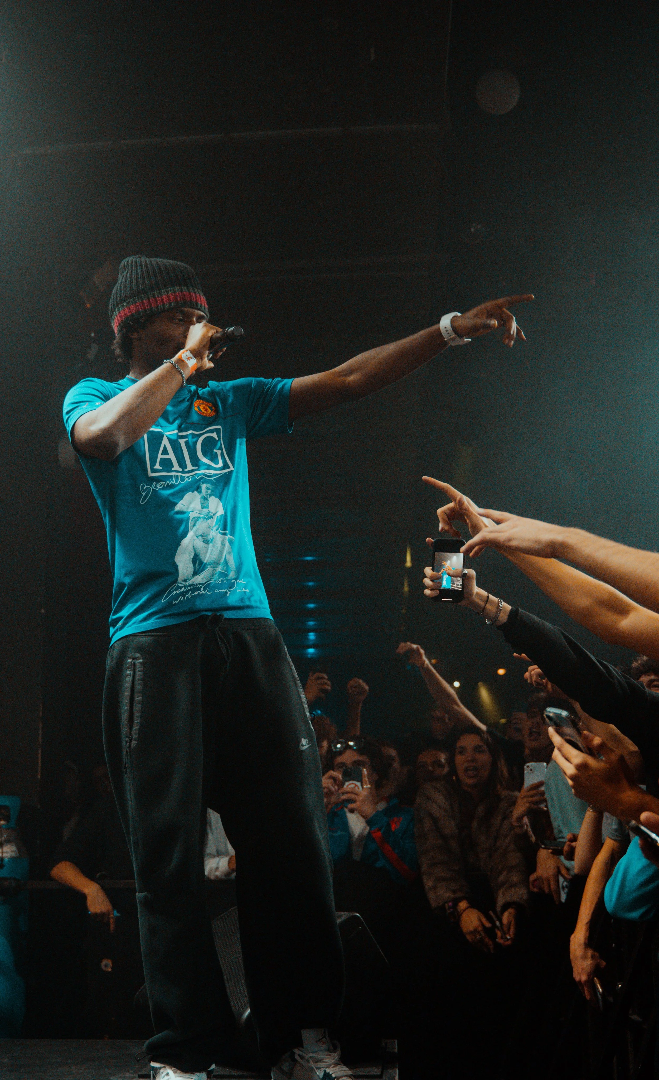 Un artiste en t-shirt bleu avec une casquette noire et rouge, tenant un micro, en train de performer sur scène devant une foule, certains spectateurs prennent des photos ou vidéos avec leurs téléphones.
