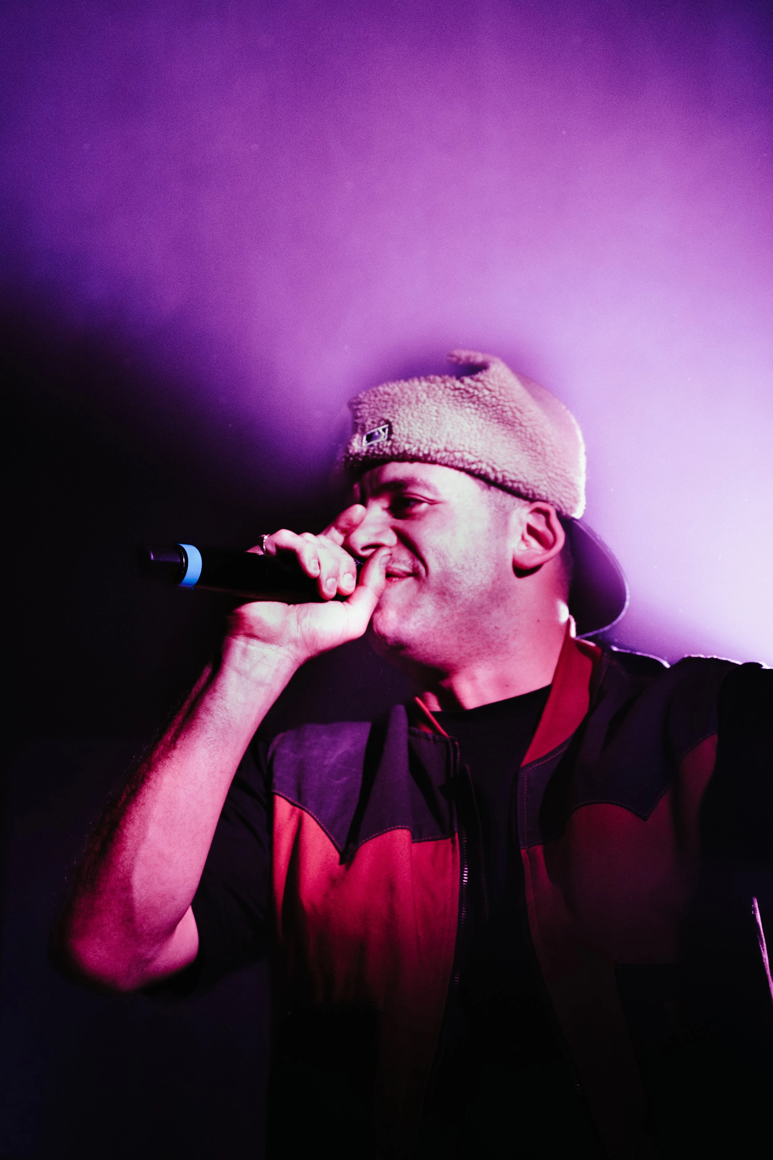 Un homme portant un bonnet en peluche, tenant un microphone, en train de chanter ou de parler lors d'un spectacle, avec un éclairage violet et sombre en arrière-plan.