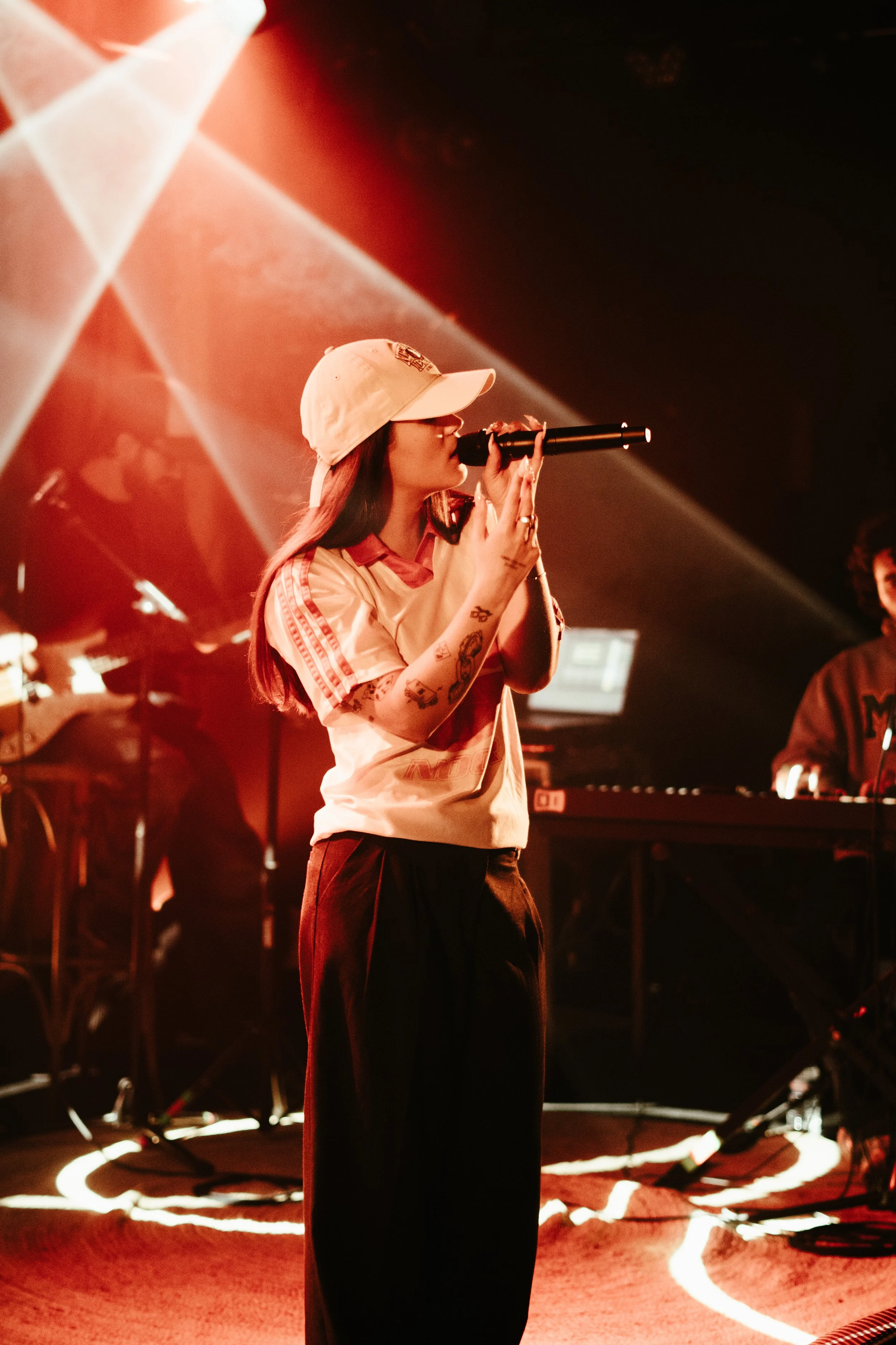 Une femme portant une casquette blanche chante dans un micro sur scène, avec un groupe de musiciens en arrière-plan, sous des lumières rouges et blanc.