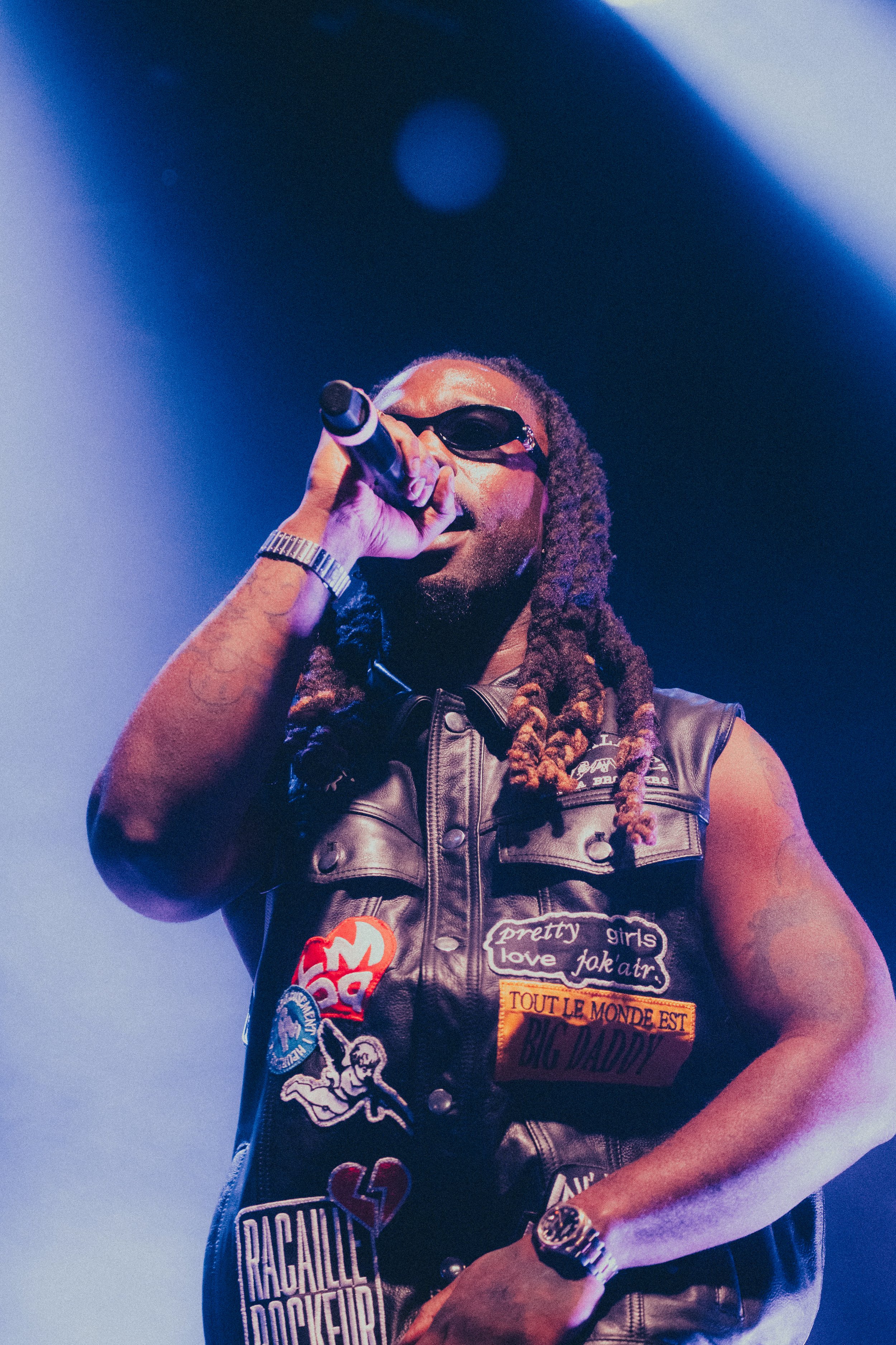 Un homme portant des lunettes de soleil, un gilet en cuir orné de patchs et de broderies, tenant un microphone en scène, sous des lumières de scène bleues et violettes.