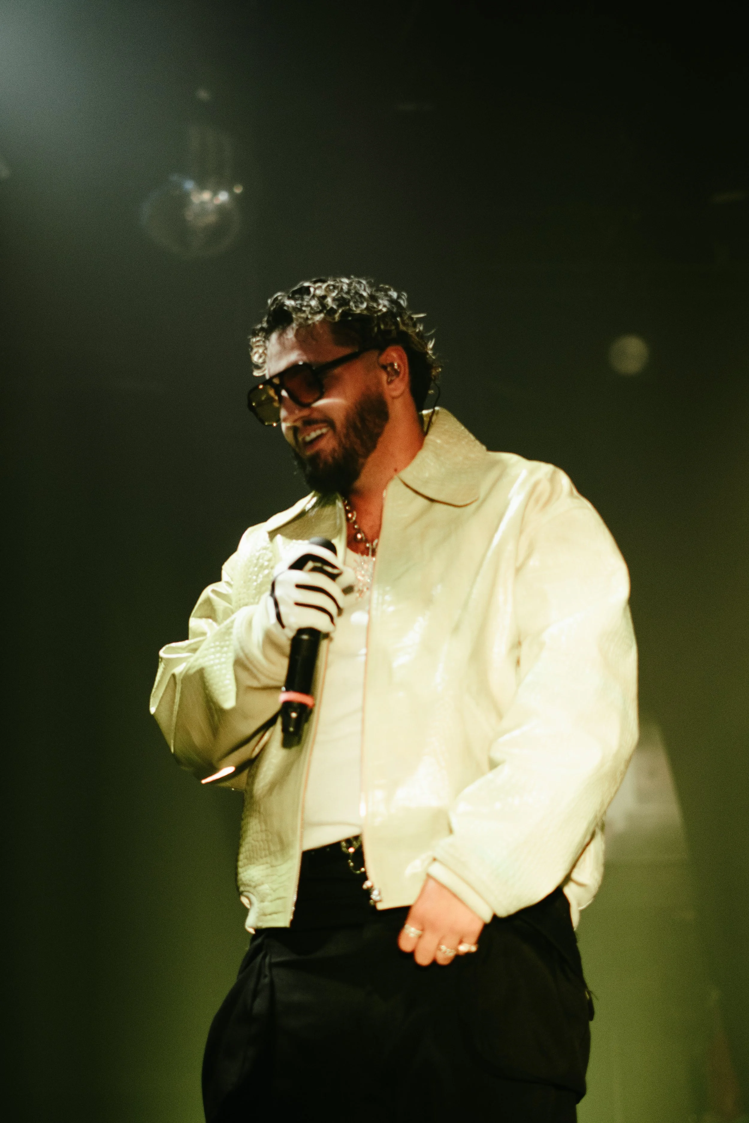 Un homme souriant portant des lunettes de soleil, une veste en satin blanc, et tenant un microphone sur scène.