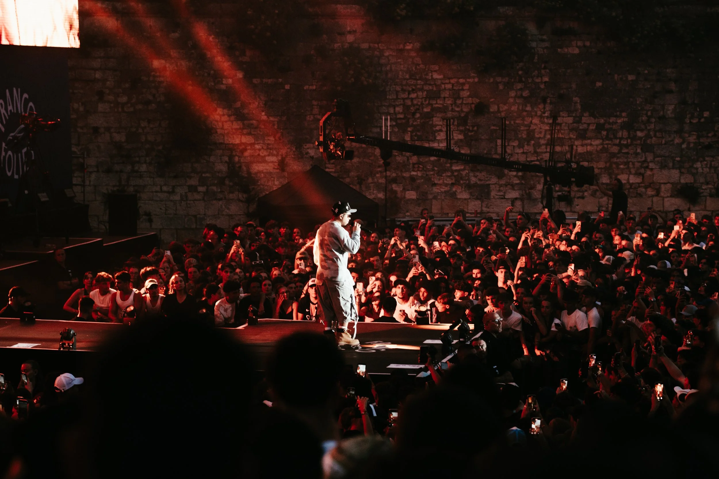 Un chanteur avec un micro sur scène lors d'un concert, avec un public dense et nombreux, éclairé par des lumières rouges, devant un mur en pierre.