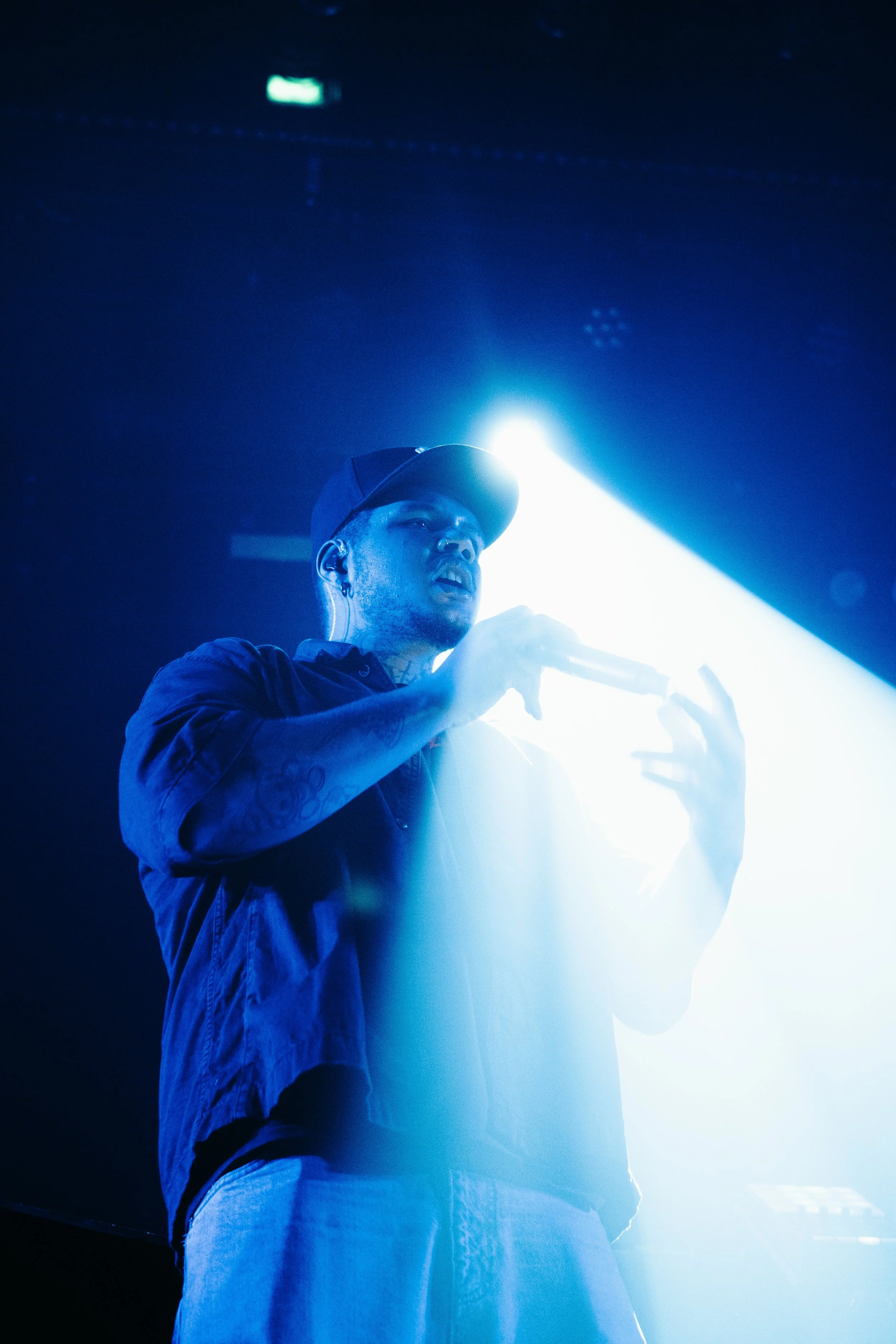 Un homme avec une casquette tient un micro sur scène avec un éclairage bleu intense derrière lui.