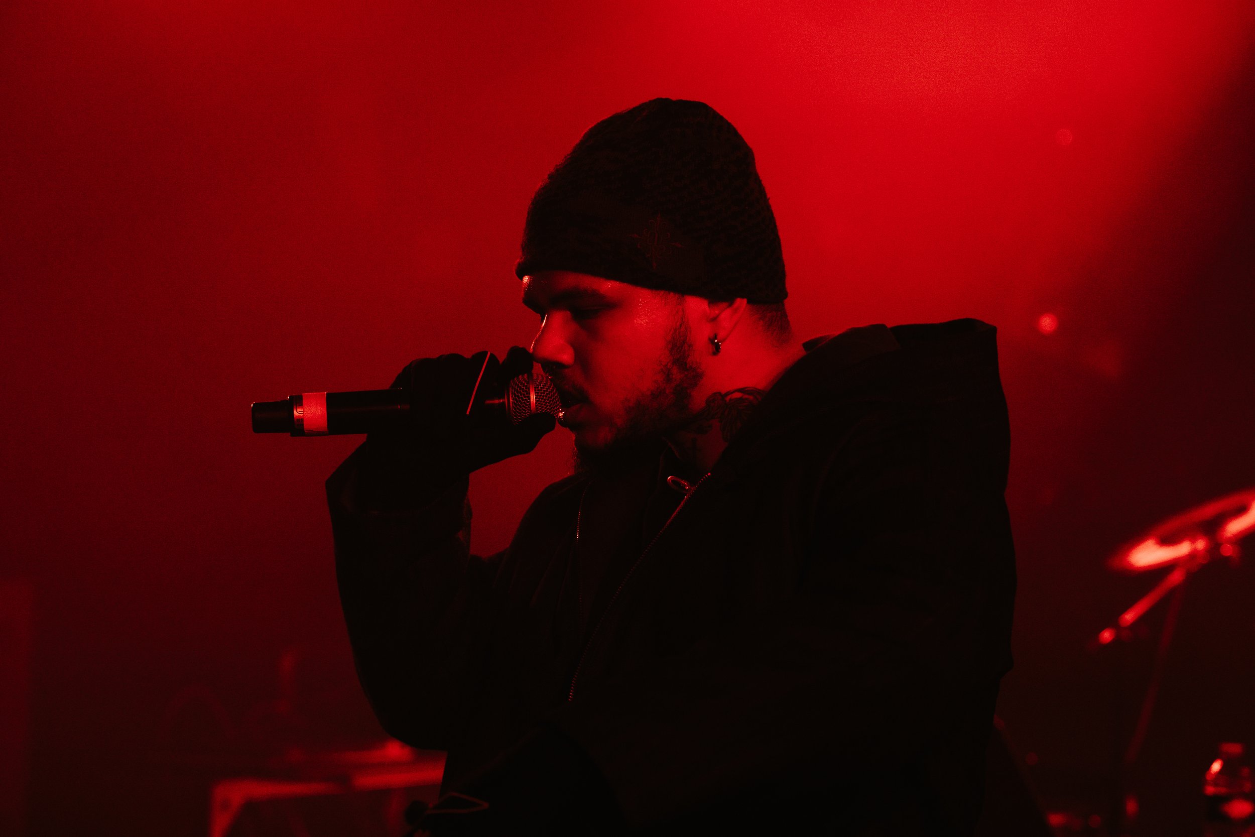 Un jeune homme chantant dans un microphone, vêtu d'une veste noire et portant un bonnet noir, éclairé par une lumière rouge dans un environnement sombre.