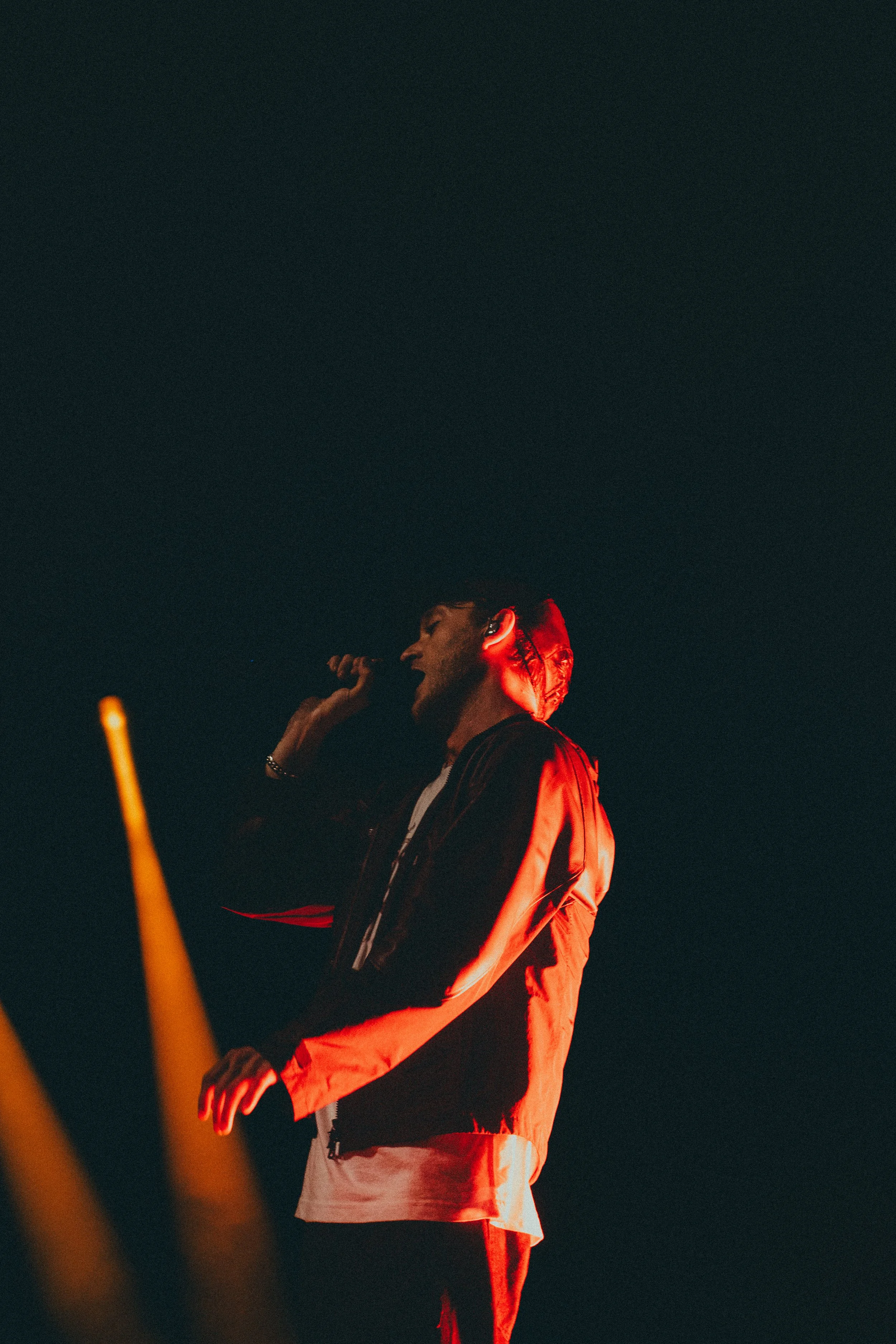 Un homme chante lors d'une performance dans un éclairage rougeâtre, portant une veste noire et un t-shirt blanc, dans un décor sombre.