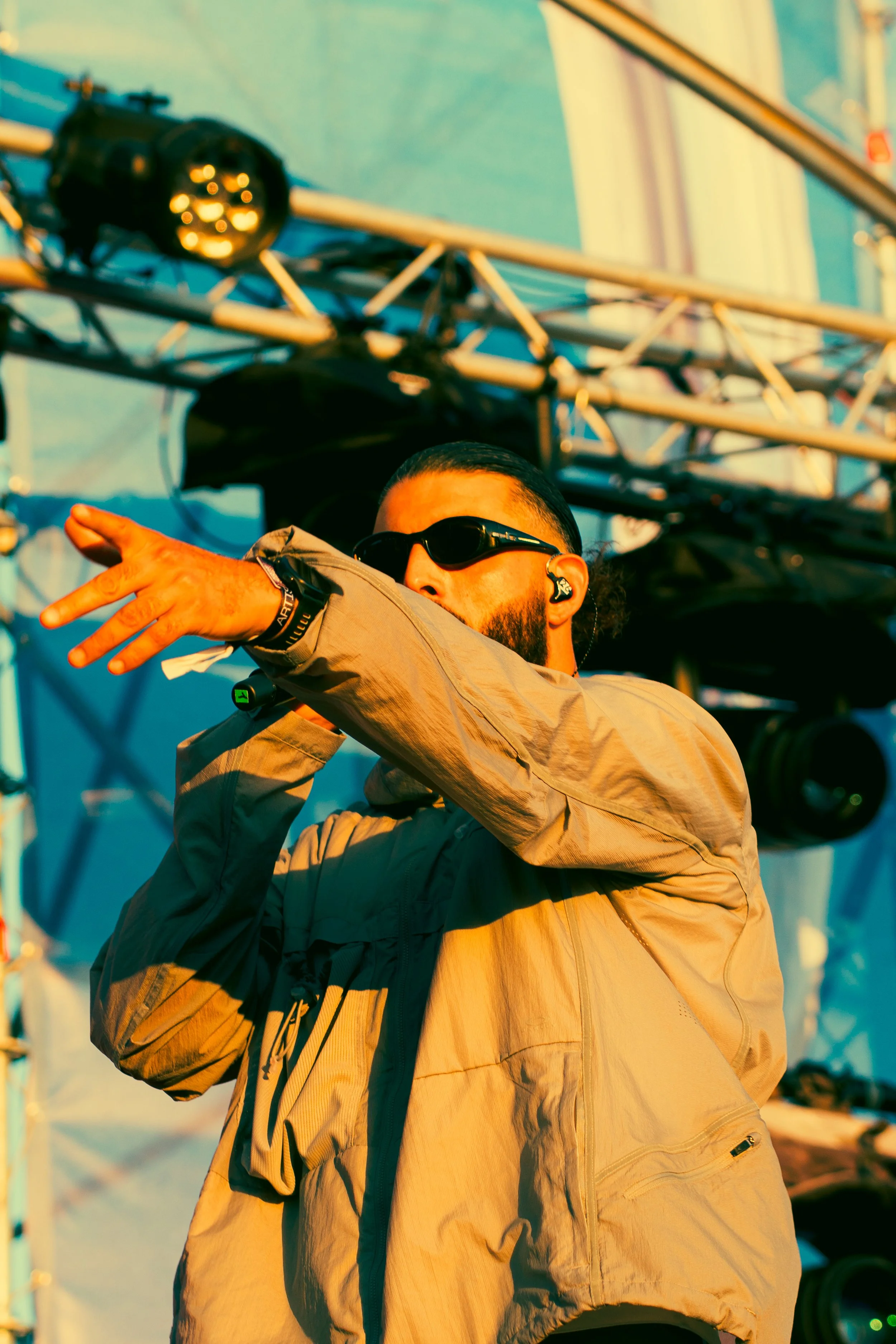 Un homme portant des lunettes de soleil, une veste beige, en train de faire un geste avec sa main lors d'un spectacle en plein air avec un équipement de sonorisation en arrière-plan.