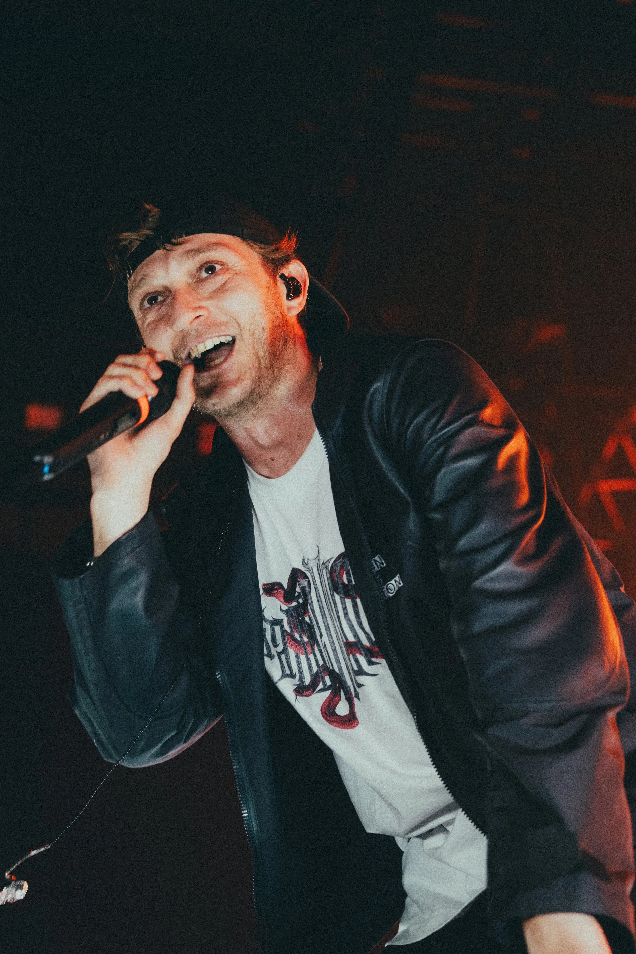 Un homme chantant en microphone lors d'un concert, portant un sweat-shirt blanc avec un design, une veste en cuir et un chapeau. ambiance de scène avec éclairage rouge/orange.