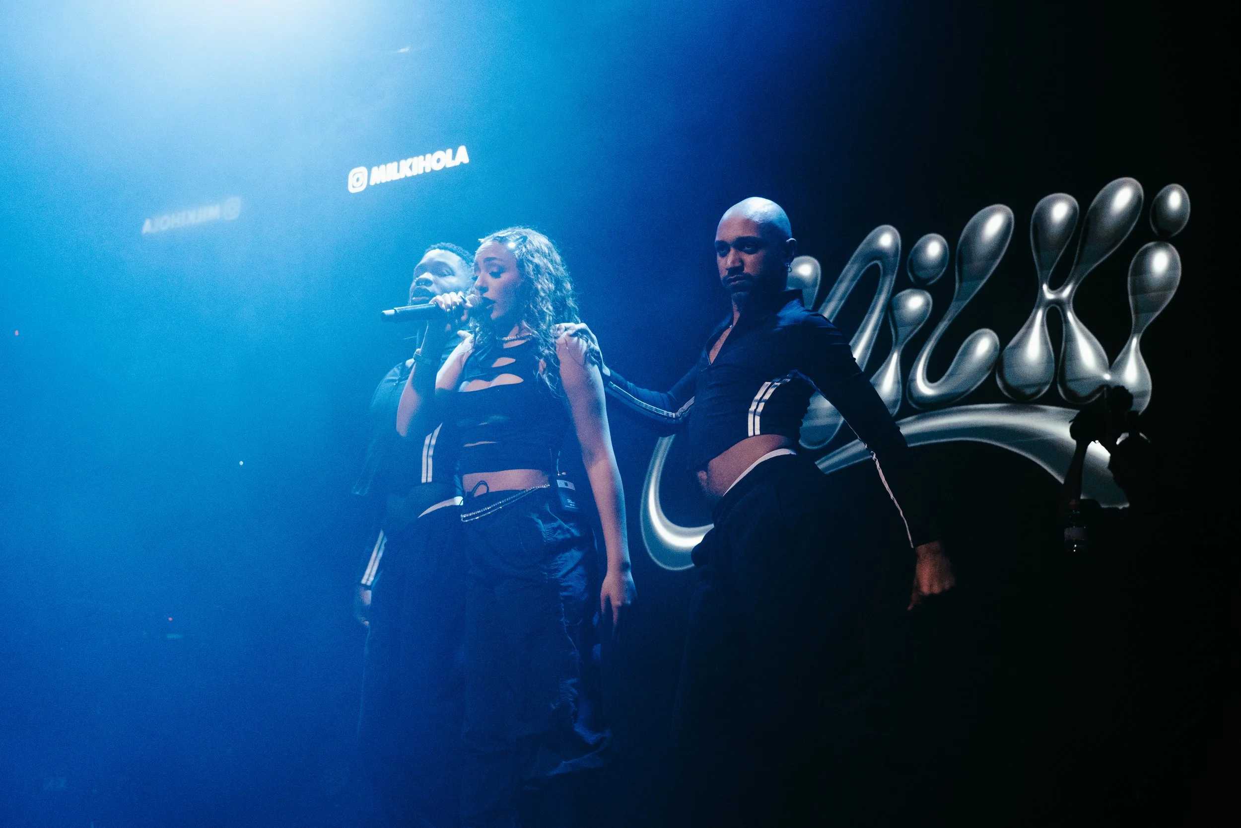 Trois personnes sur scène, deux femmes et un homme, dans un éclairage bleu, avec un logo en arrière-plan.