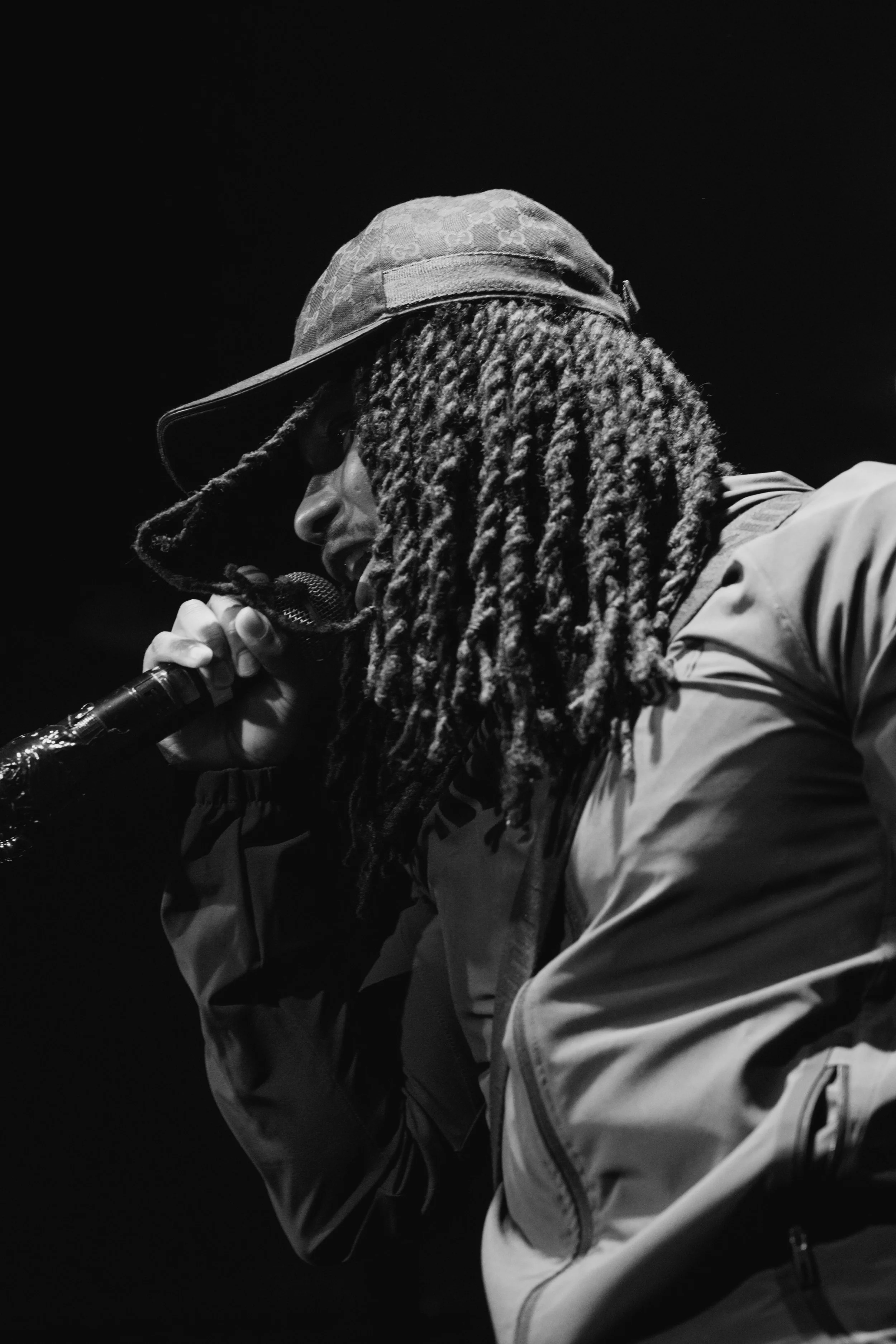Une personne avec des dreadlocks portant un chapeau et une veste, tenant un microphone, en train de chanter ou parler, photographiée en noir et blanc.