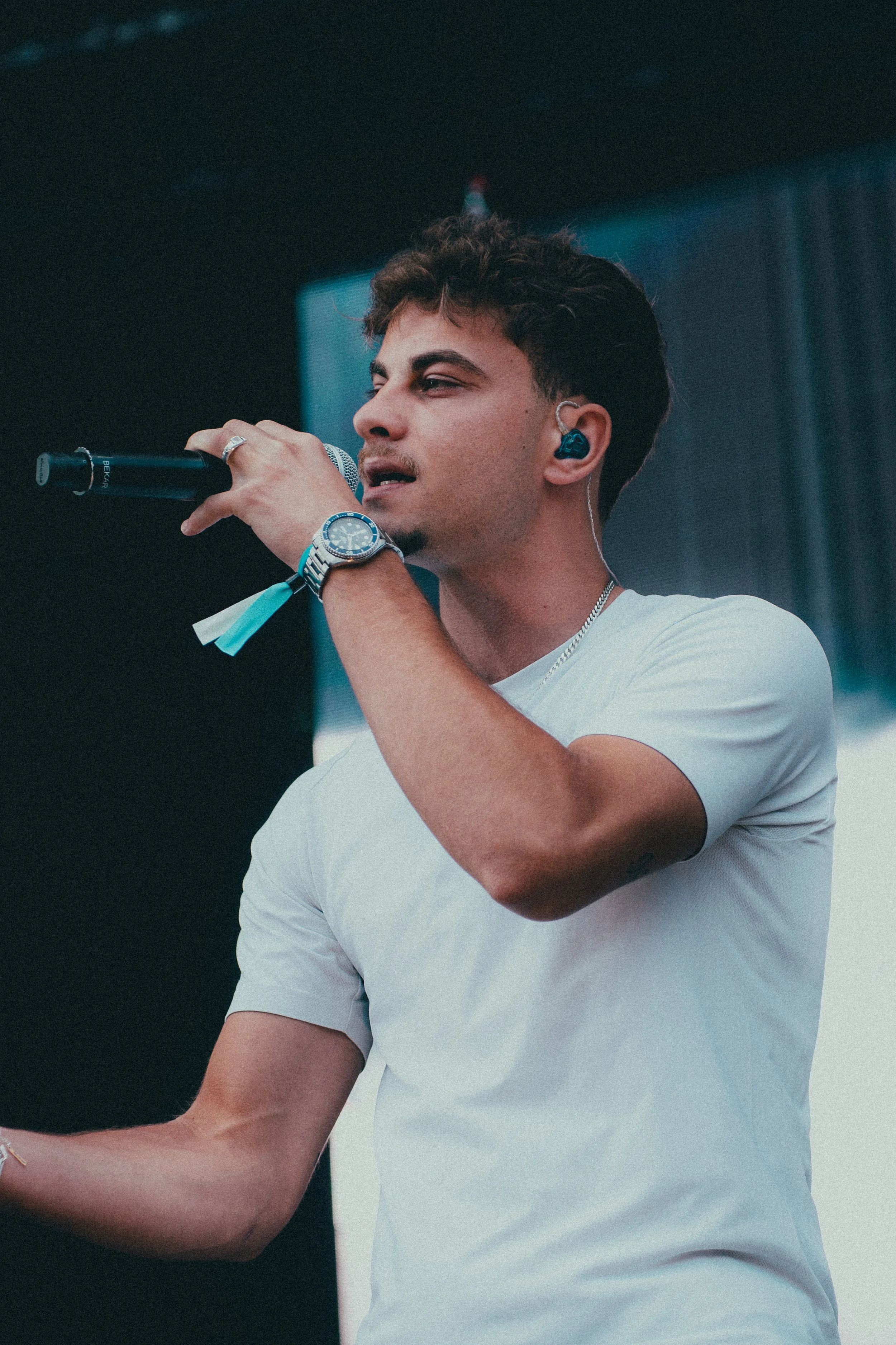 Jeune homme chantant en microphone lors d'un concert, portant un t-shirt blanc, une montre et un écouteur bleu.