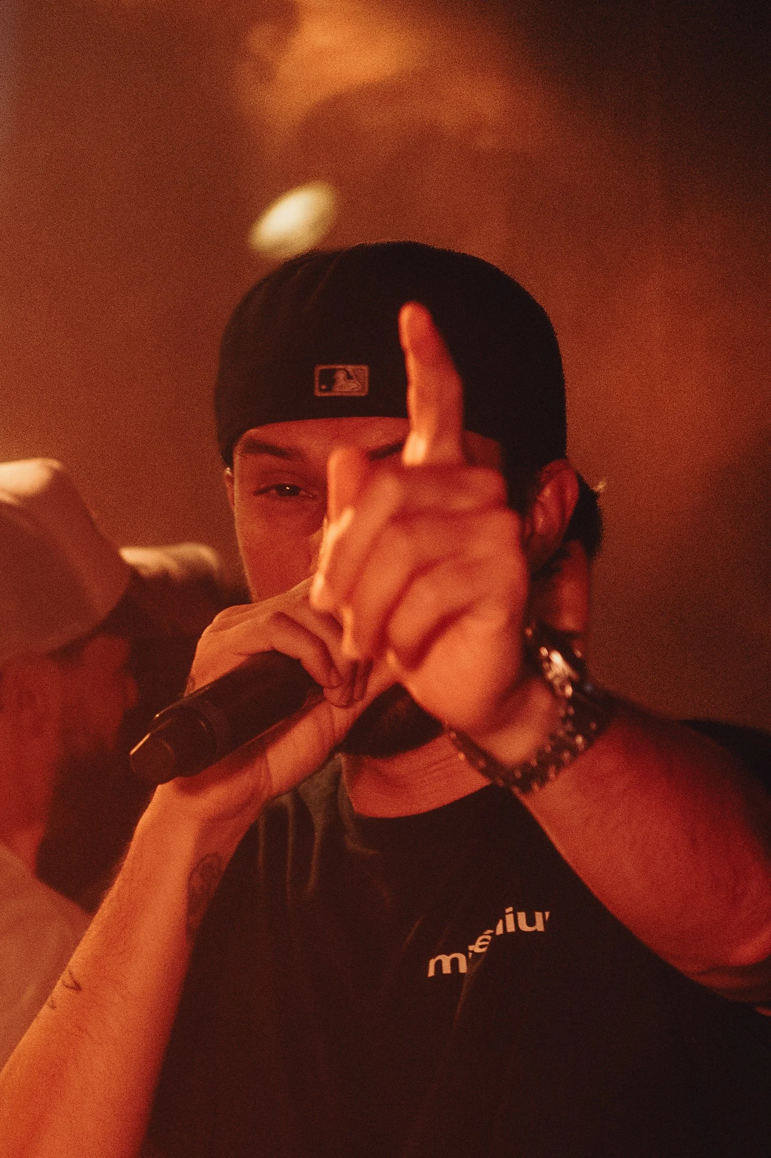 Un jeune homme portant une casquette noire, portant un bracelet en chaîne et un t-shirt noir, regarde la caméra tout en faisant un geste avec sa main. La photo a une ambiance sombre avec un arrière-plan rougeâtre.