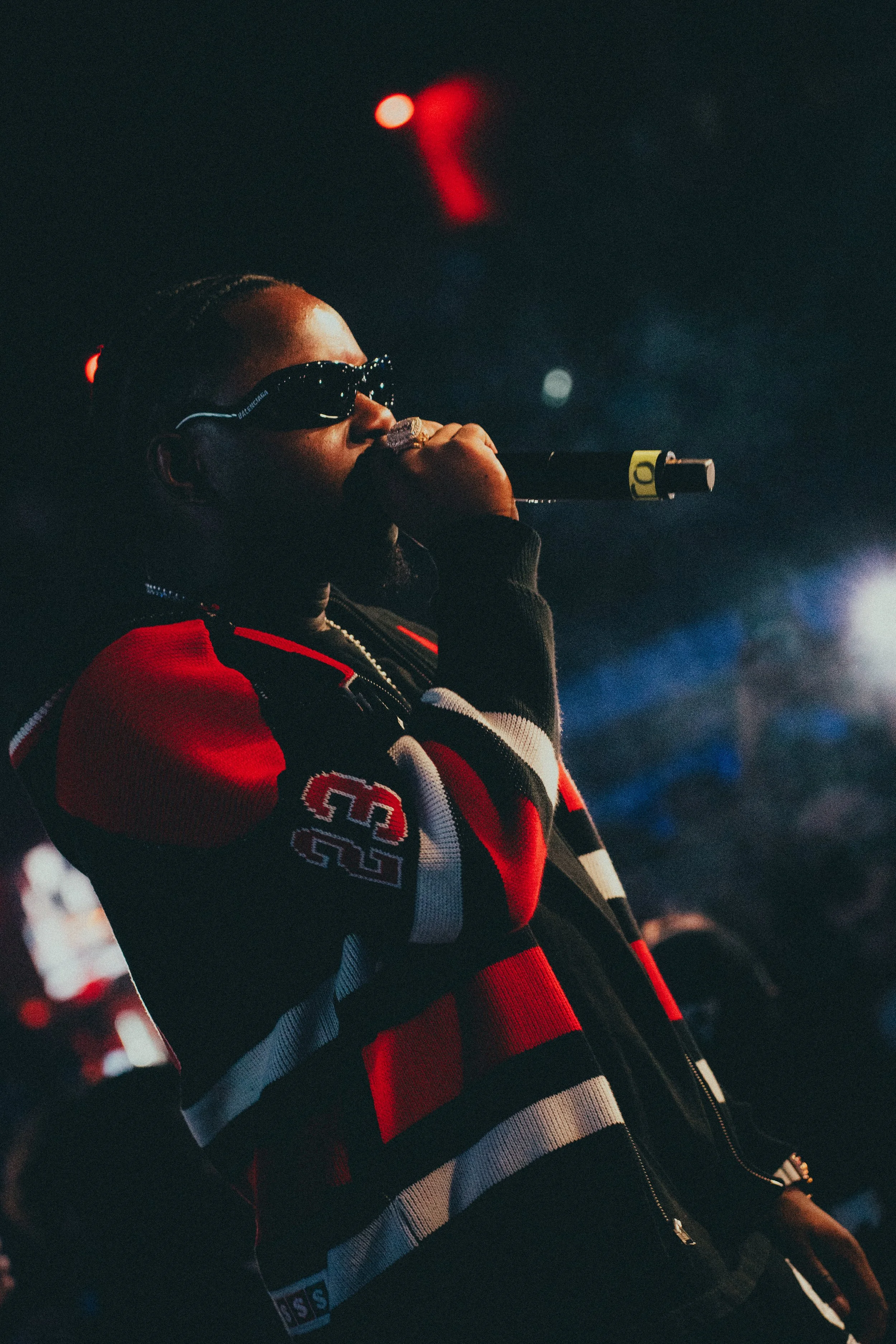 Une personne portant des lunettes de soleil noires, tenant un microphone, portant un sweat à capuche avec des couleurs rouge, noir et blanc lors d'une performance sur scène dans une ambiance sombre avec des lumières colorées.