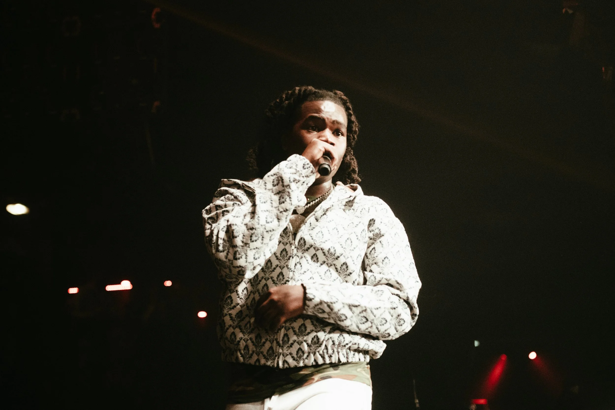 Une personne chante en tenant un micro sur scène, vêtue d'un sweat à motifs et d'un pantalon en camouflage, sous des lumières de scène.