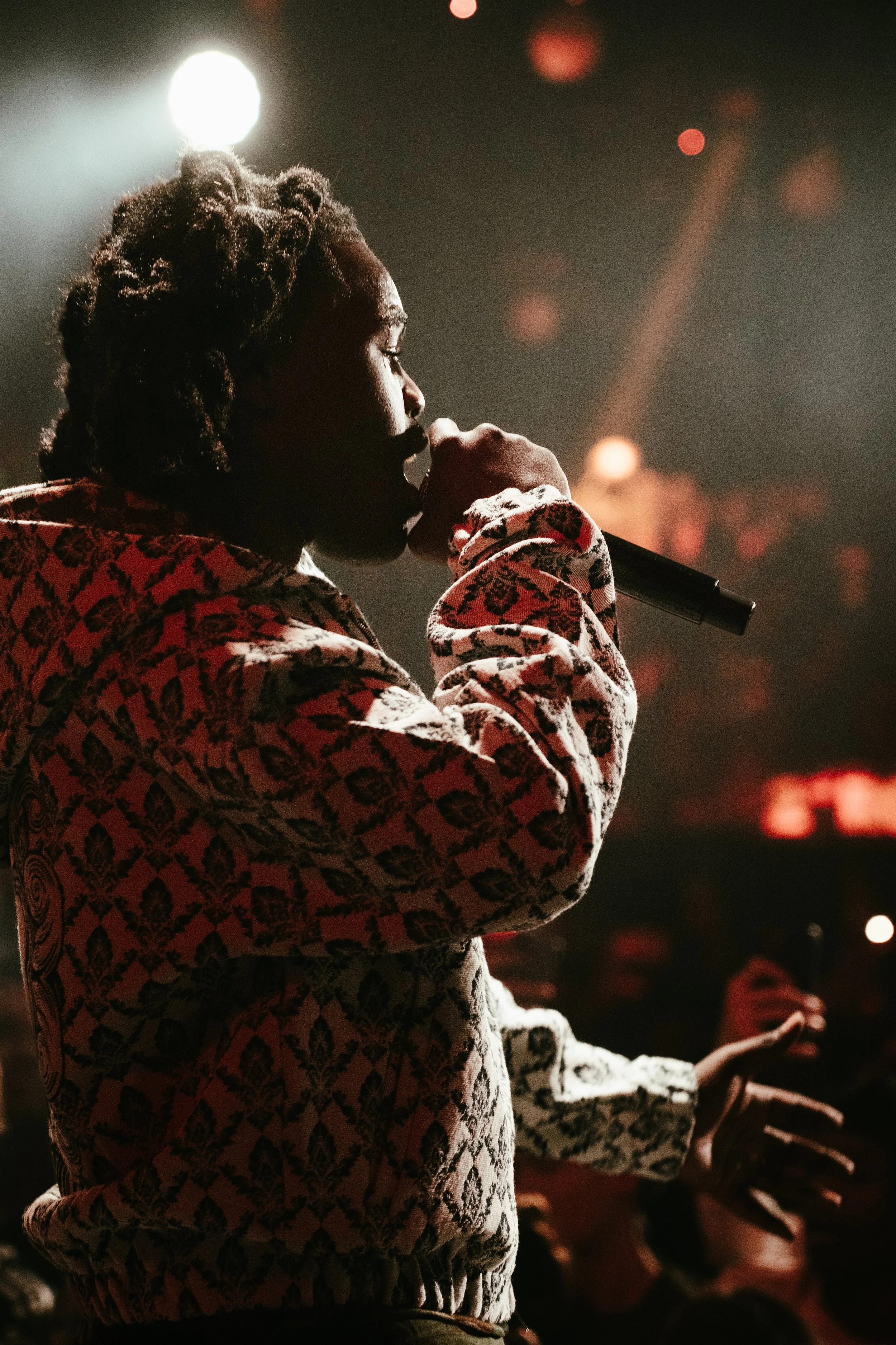 Une personne chantant ou parlant dans un micro lors d'une performance sur scène, avec un éclairage dramatique et un arrière-plan sombre.