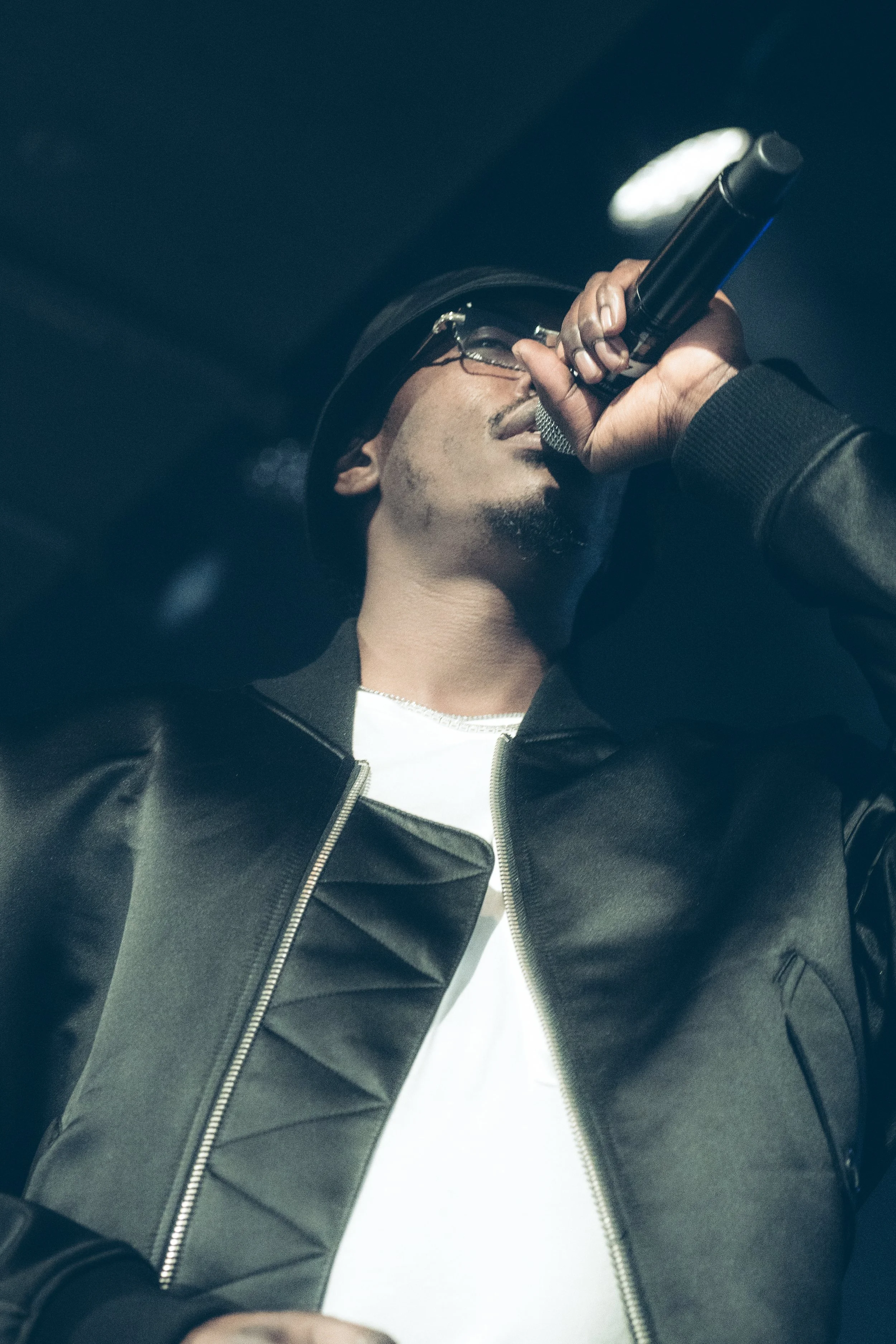 Un homme chante avec un microphone, portant un chapeau, des lunettes et une veste en cuir noir.