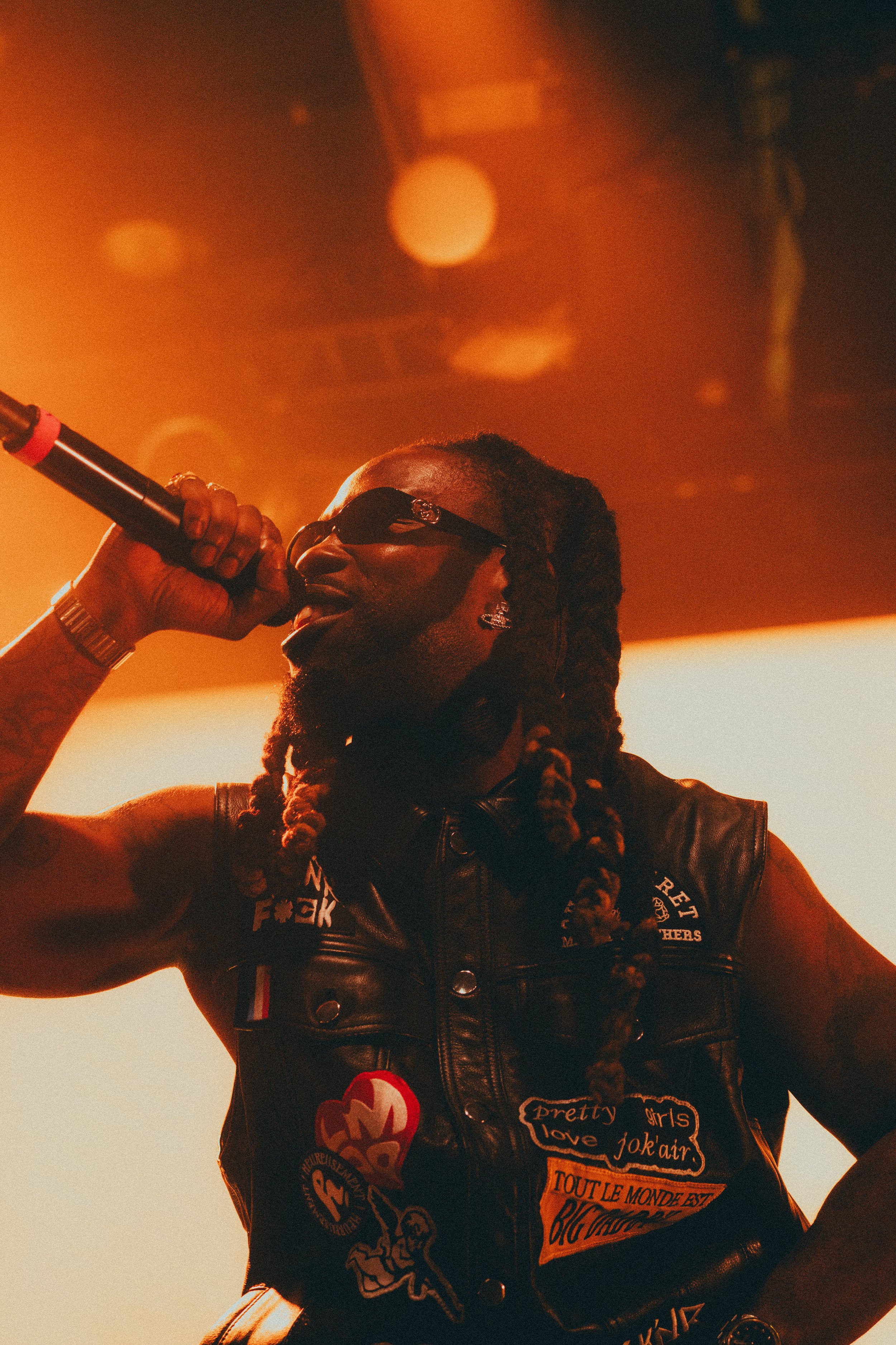 Un homme portant des lunettes de soleil noires, une veste en cuir avec des patches, tenant un microphone en scène lors d'une performance musicale, sous un éclairage chaud et brumeux.
