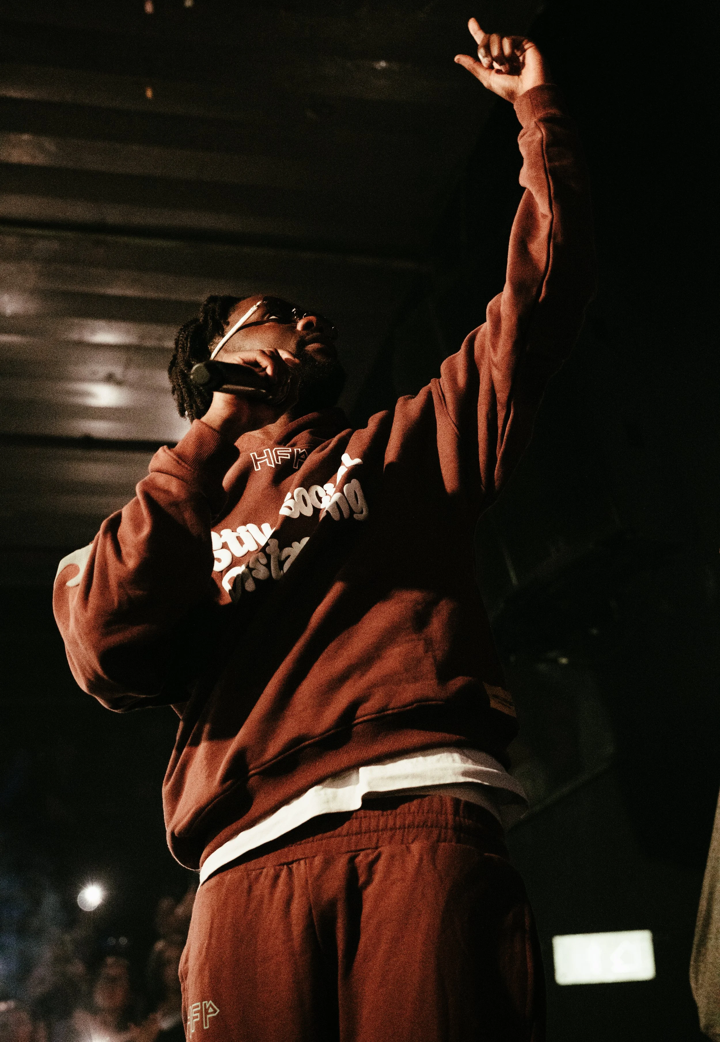 Un homme portant une veste marron et des lunettes tient un microphone et fait un geste avec la main levée dans un environnement sombre, probablement lors d'une performance ou d'un discours.