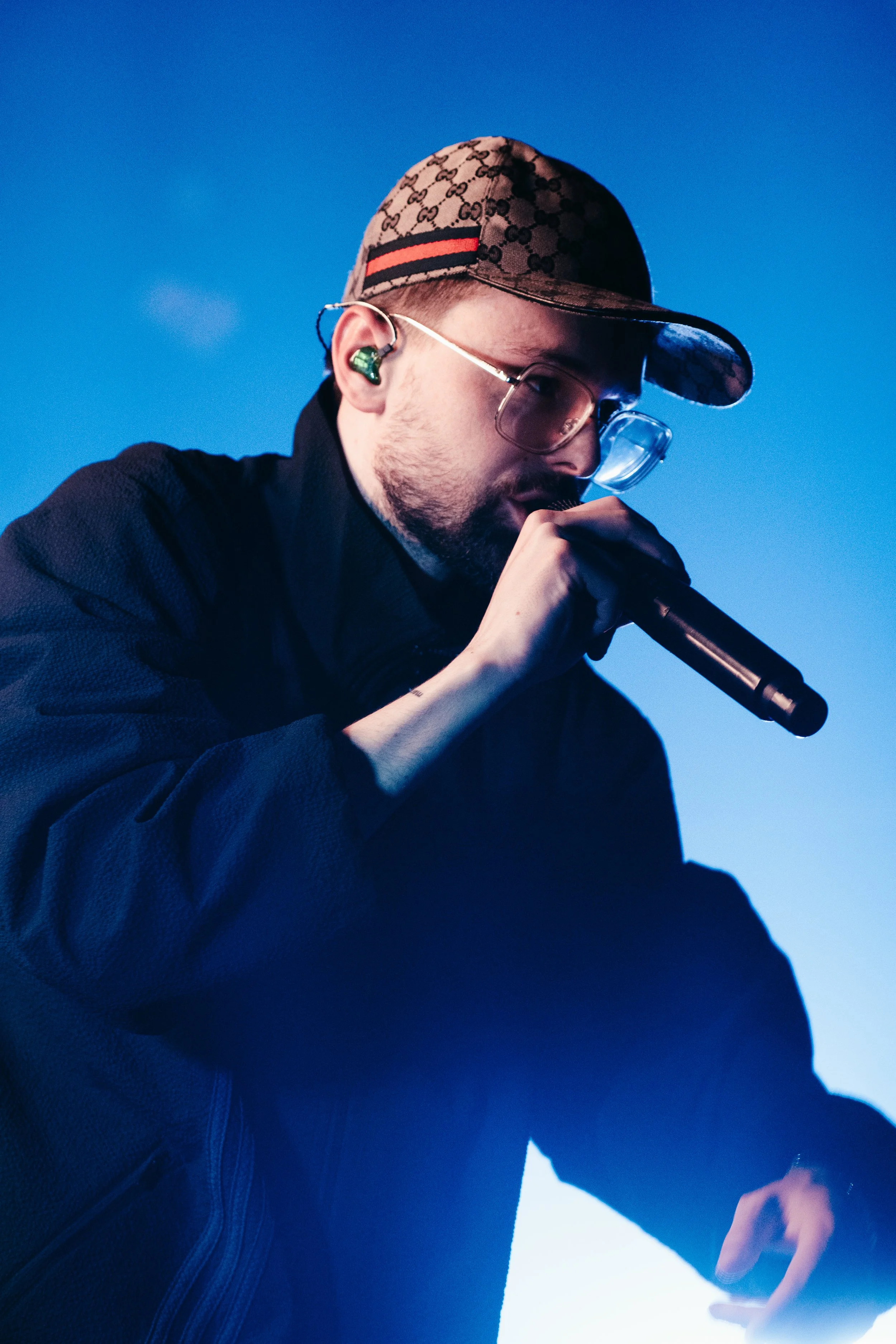 Un homme chante ou rappe en studio, portant un casque Gucci, des lunettes et une veste noire, avec un fond bleu.