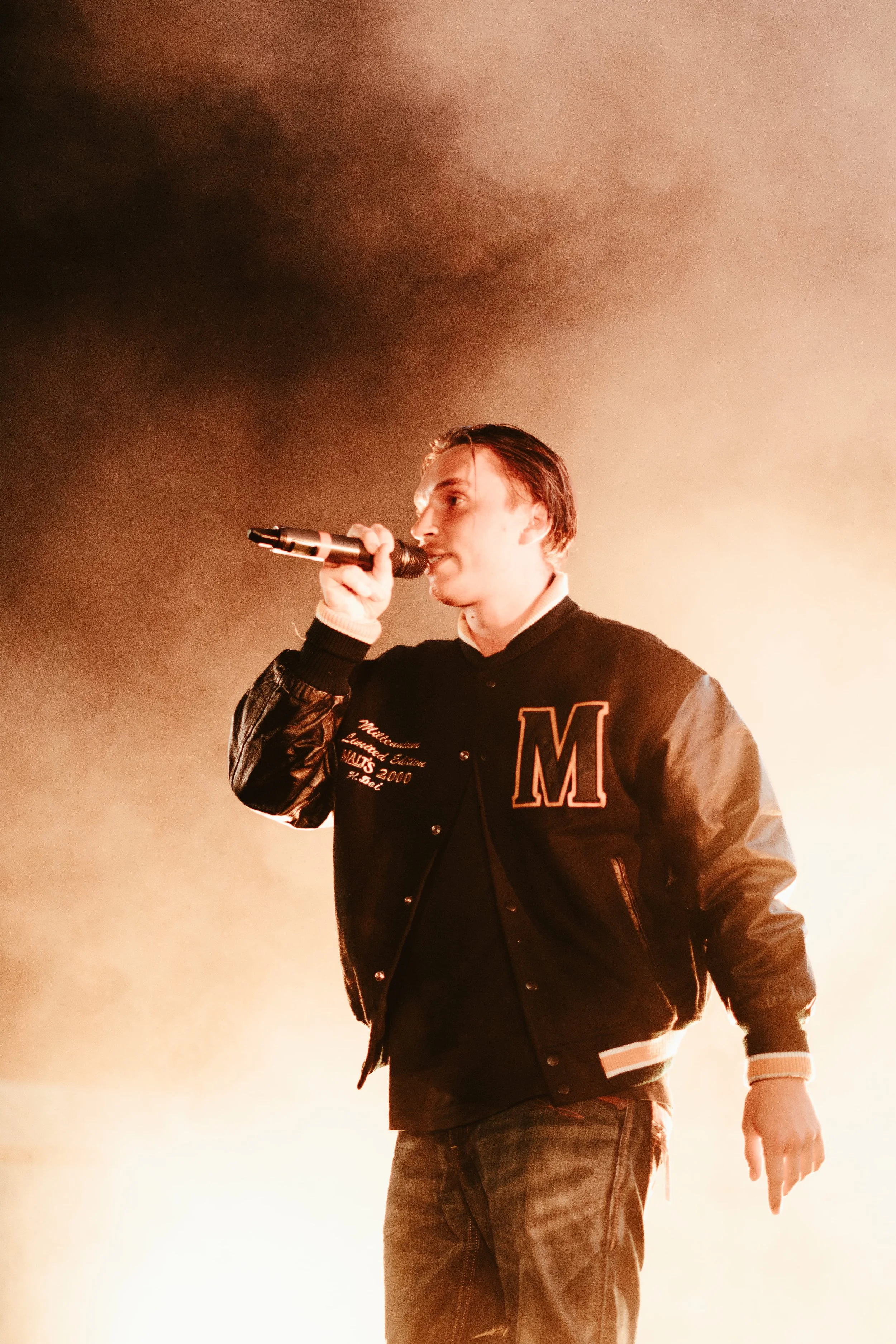 Un jeune homme chante en tenant un microphone lors d'une performance sur scène, entouré de fumée et d'une lumière chaude.