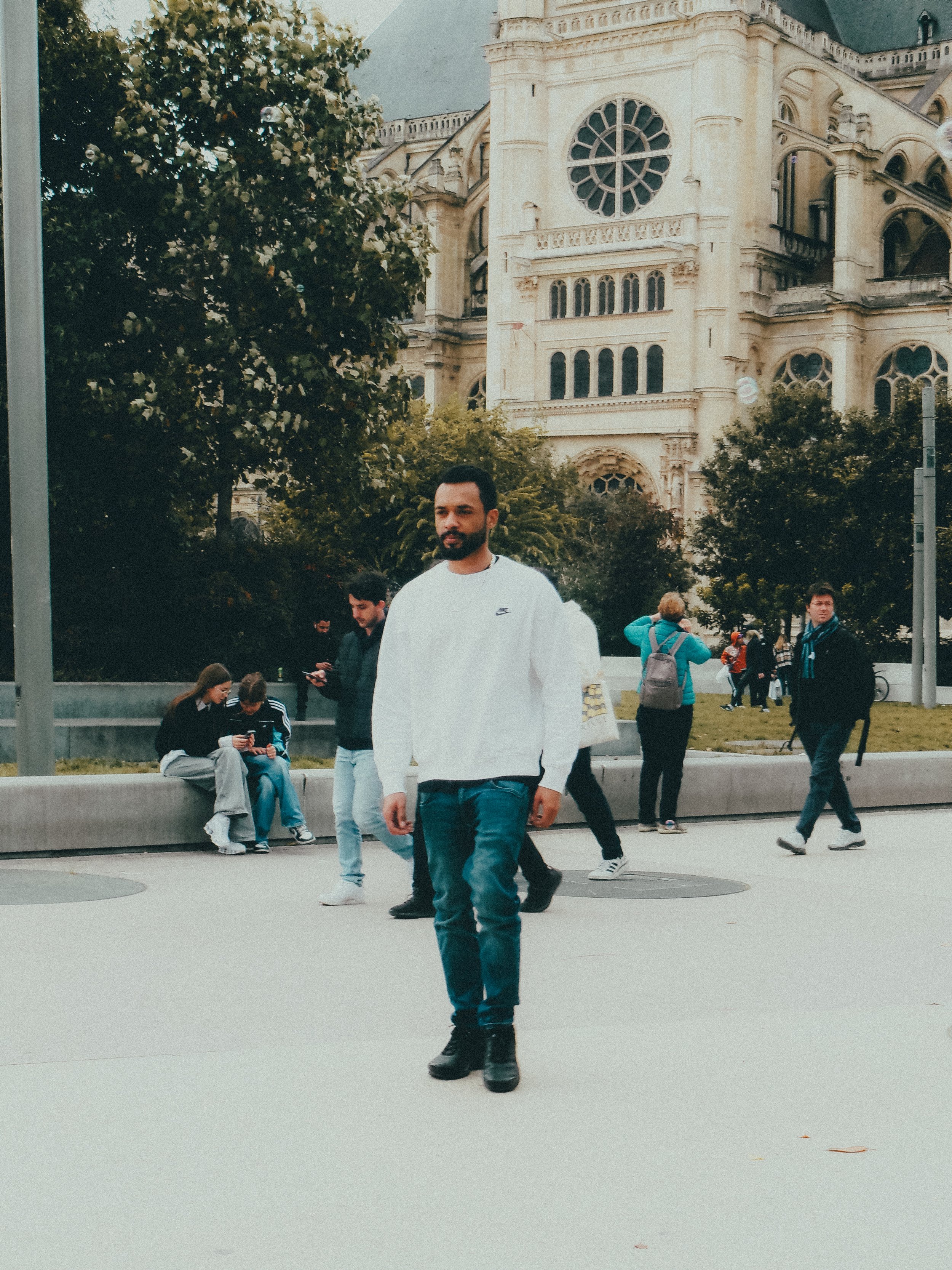 Un homme marche dans une zone urbaine devant une vieille cathédrale gothique, entouré de plusieurs autres personnes, certains assis et d'autres marchant, avec des arbres et un ciel nuageux en arrière-plan.