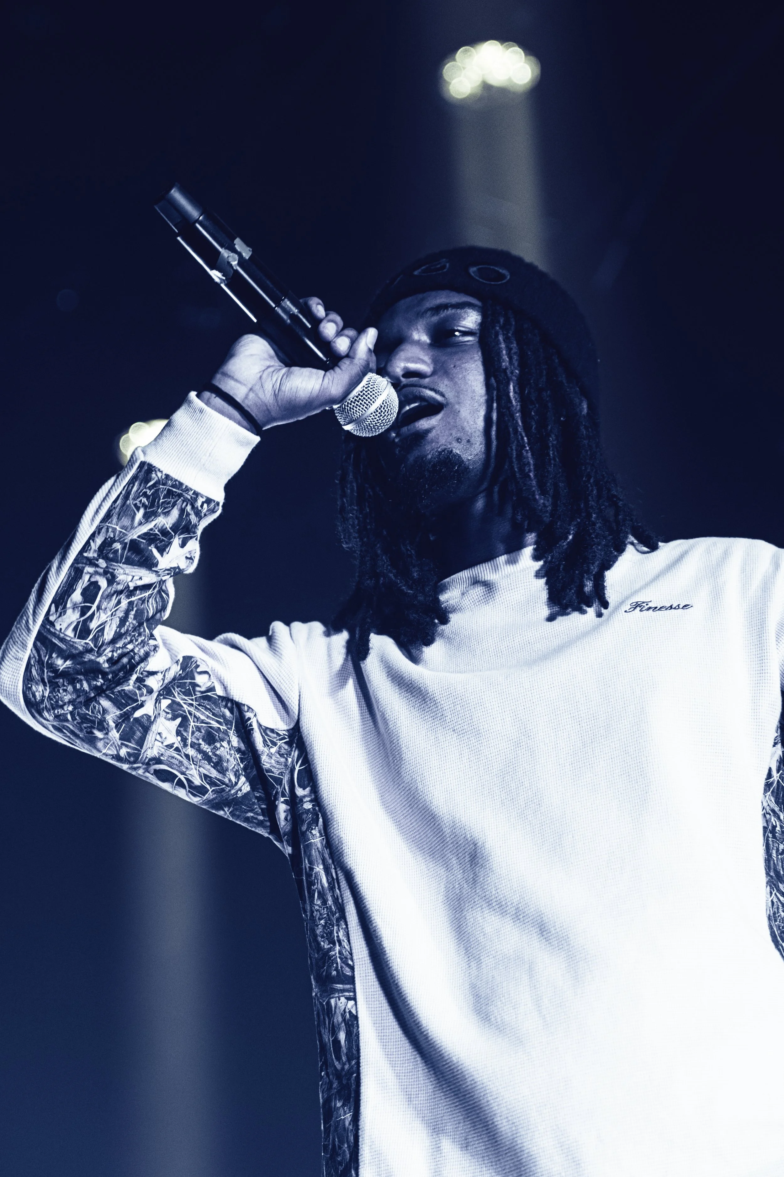 Un homme noir avec des dreadlocks, portant un sweat blanc avec des motifs sur les manches, chante dans un microphone sur scène.