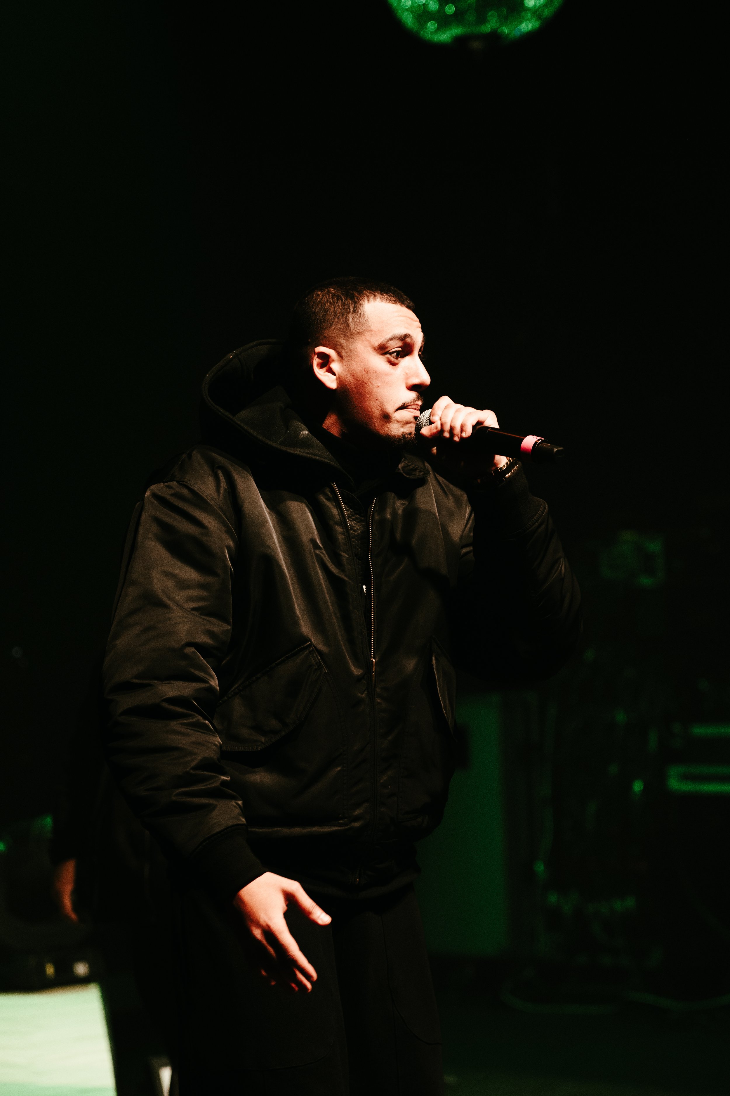 Homme avec une veste noire tenant un microphone sur scène, l'air concentré, dans une ambiance sombre avec des lumières vertes en arrière-plan.