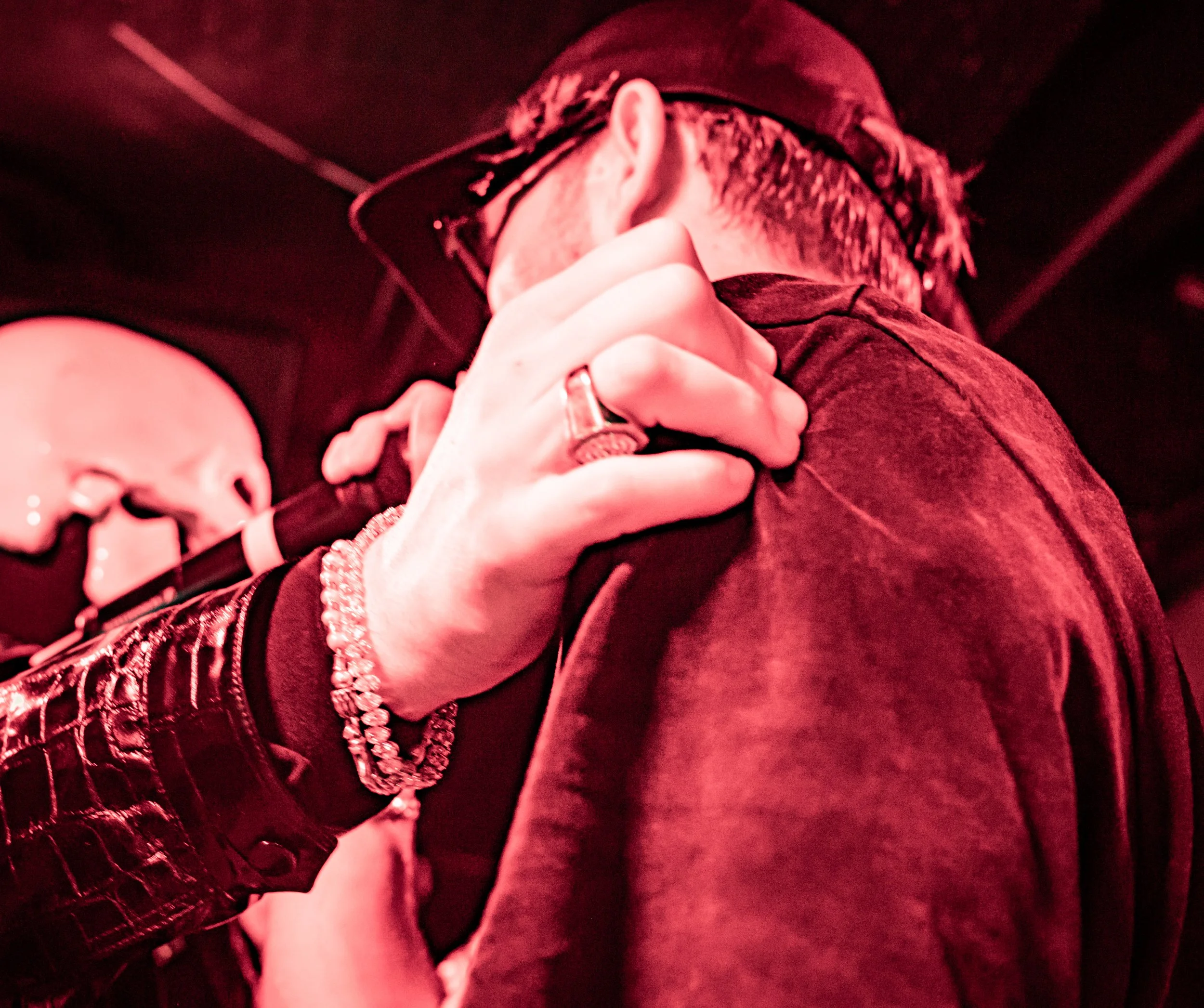 Un homme portant une casquette et des lunettes, vu de dos, en train de tenir un microphone, avec une main portée à son épaule. La scène est éclairée en rouge.