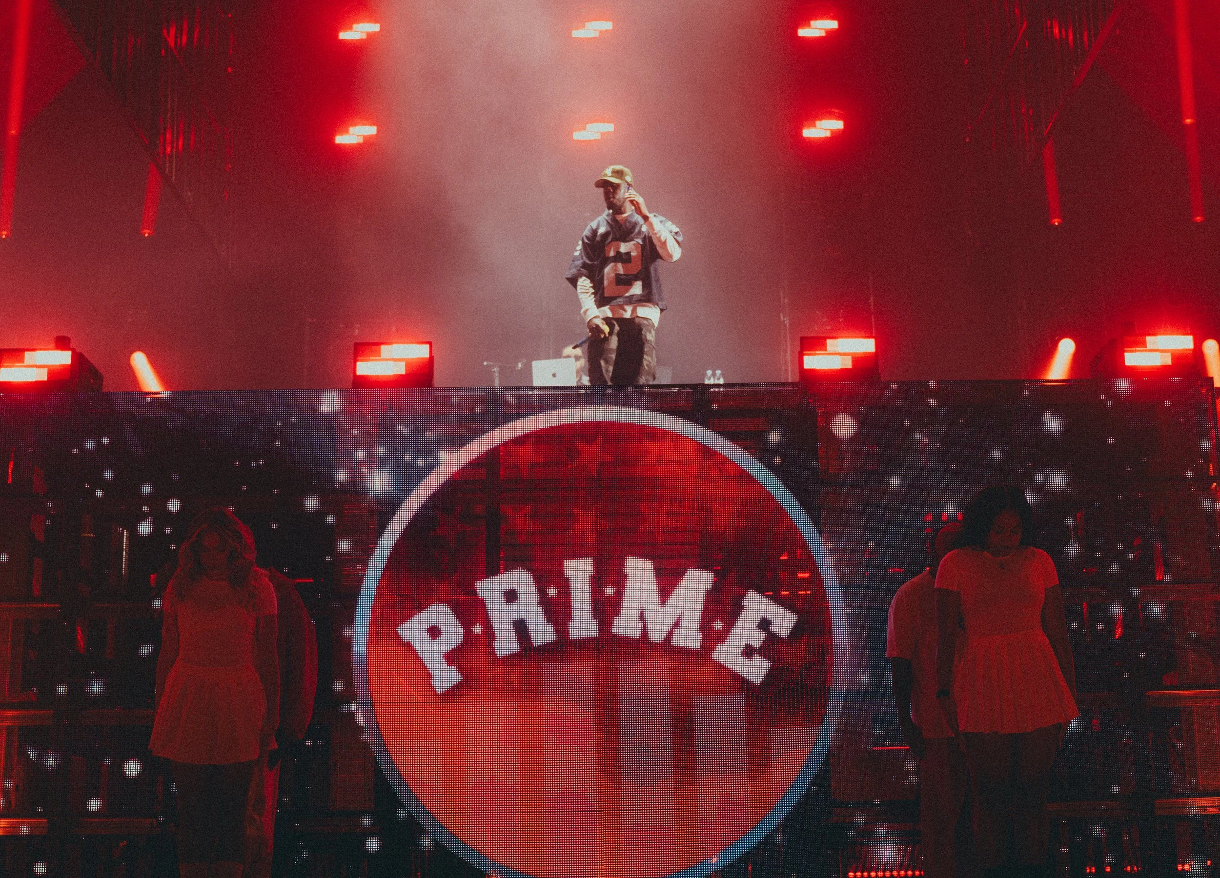 Un DJ ou un artiste performe sur scène avec un éclairage rouge, entouré de deux danseurs en arrière-plan, devant un grand écran LED affichant le logo 'PRIME'. 