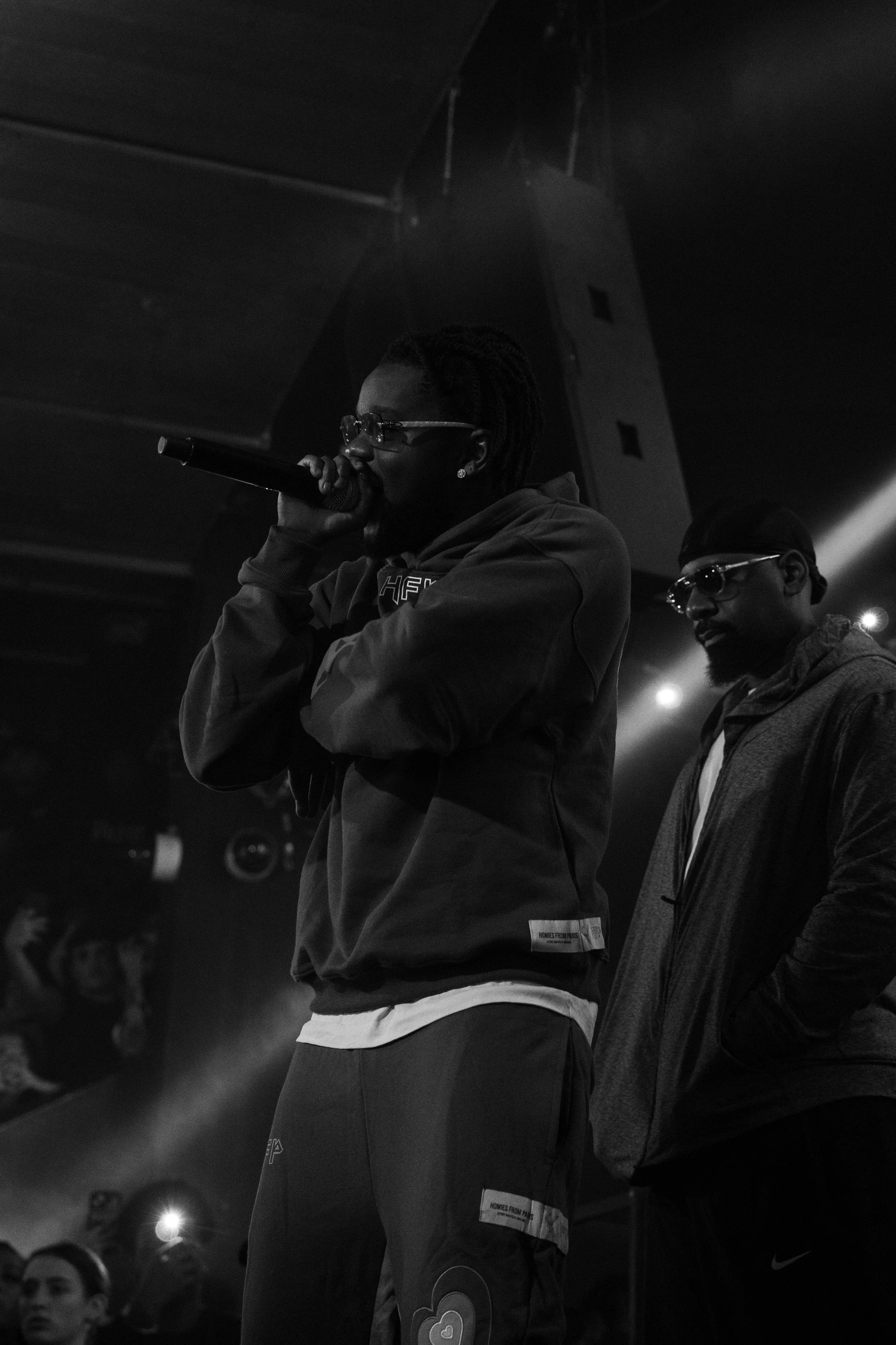 Deux hommes sur scène, l'un avec un micro, portant des lunettes de vue et un sweat à capuche, l'autre avec des lunettes de soleil et un blouson, dans une ambiance sombre avec un public en arrière-plan.