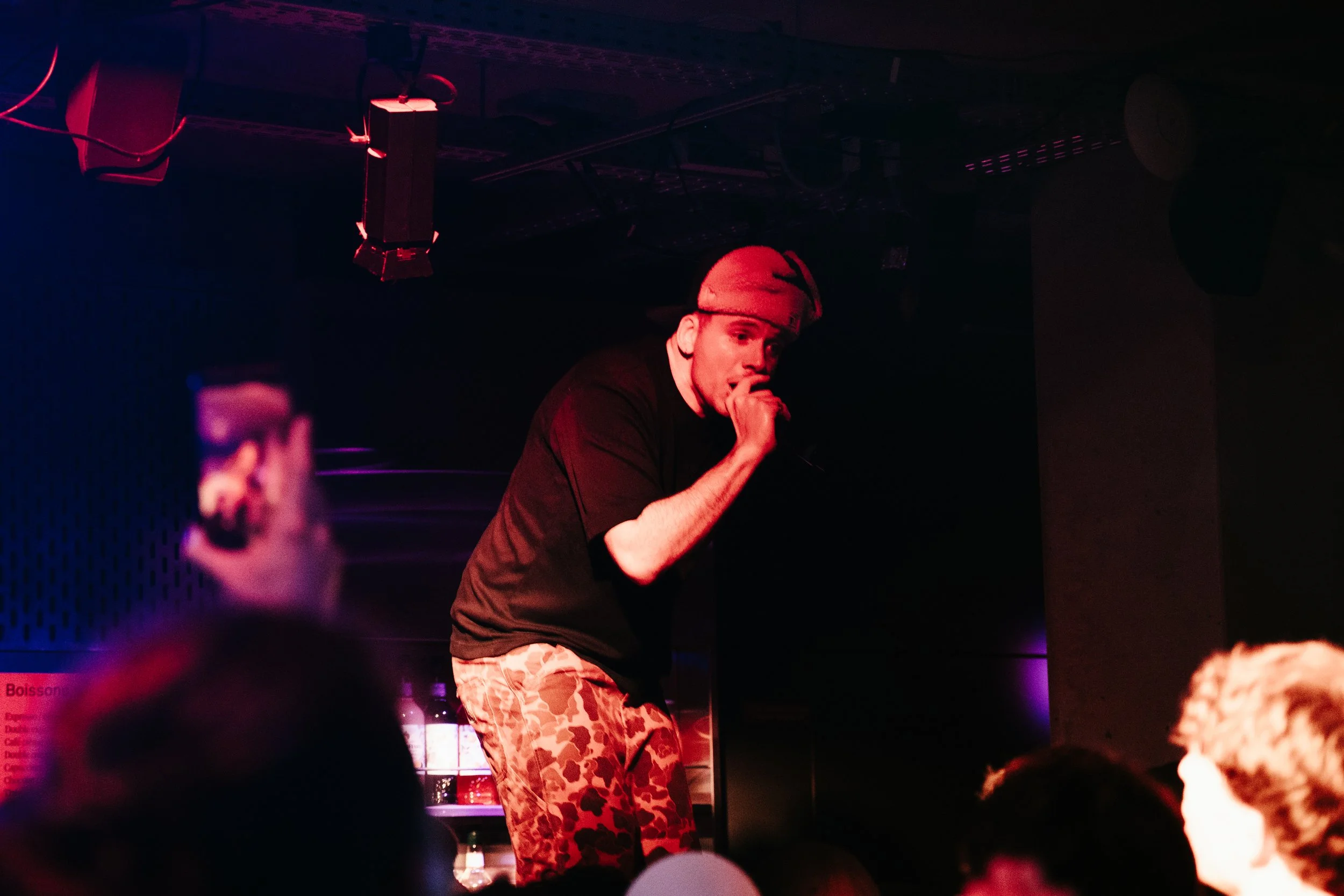 Un homme portant un chapeau beige, chantant ou parlant dans un micro sur scène, vêtu d'un t-shirt noir et pantalons camouflage, dans un environnement sombre avec des lumières rouges et violettes.