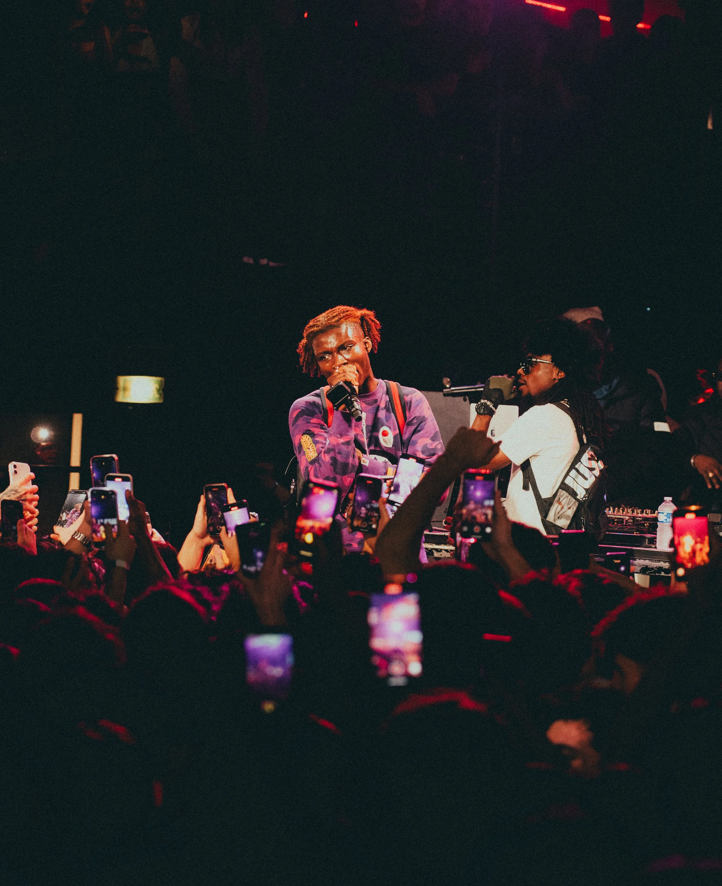 Deux artistes sur scène, un chantant dans un micro et l'autre contrôlant la musique, entourés de nombreux spectateurs prenant des photos avec leurs téléphones, dans une ambiance sombre avec des lumières rouges.