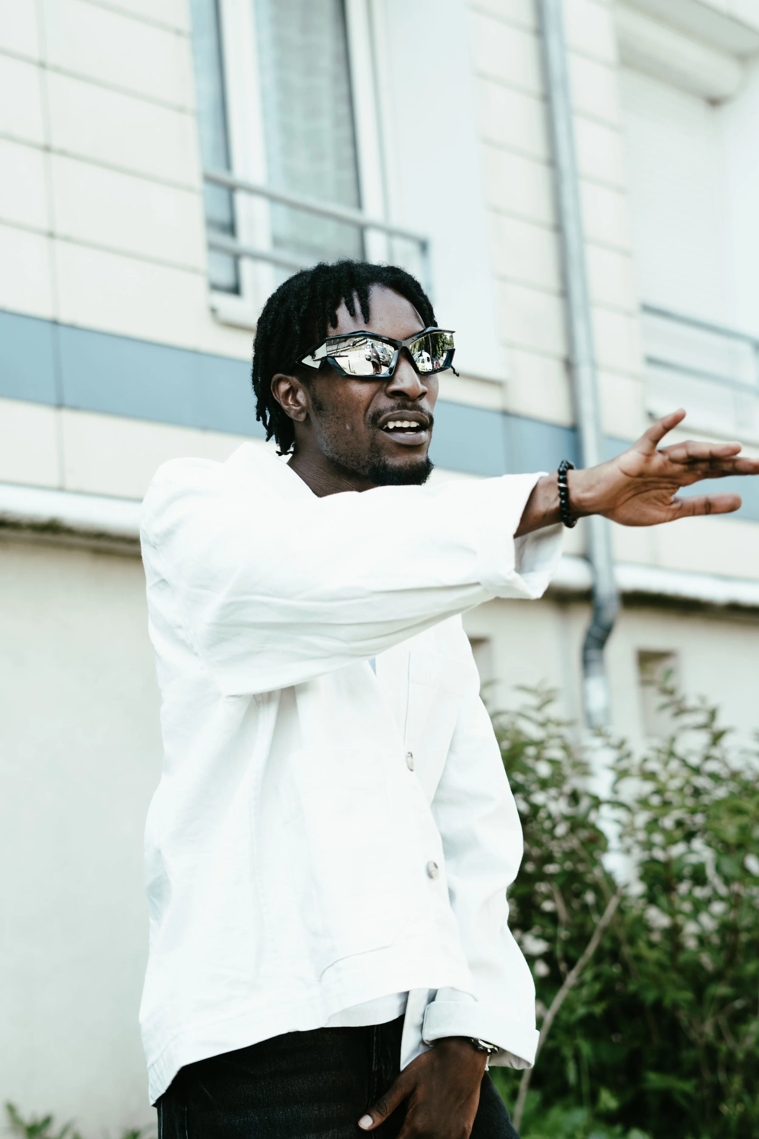 Un homme portant des lunettes de soleil réfléchissantes, une chemise blanche, et un bracelet, qui semble faire un geste ou parler, à l'extérieur devant une maison avec des fenêtres.