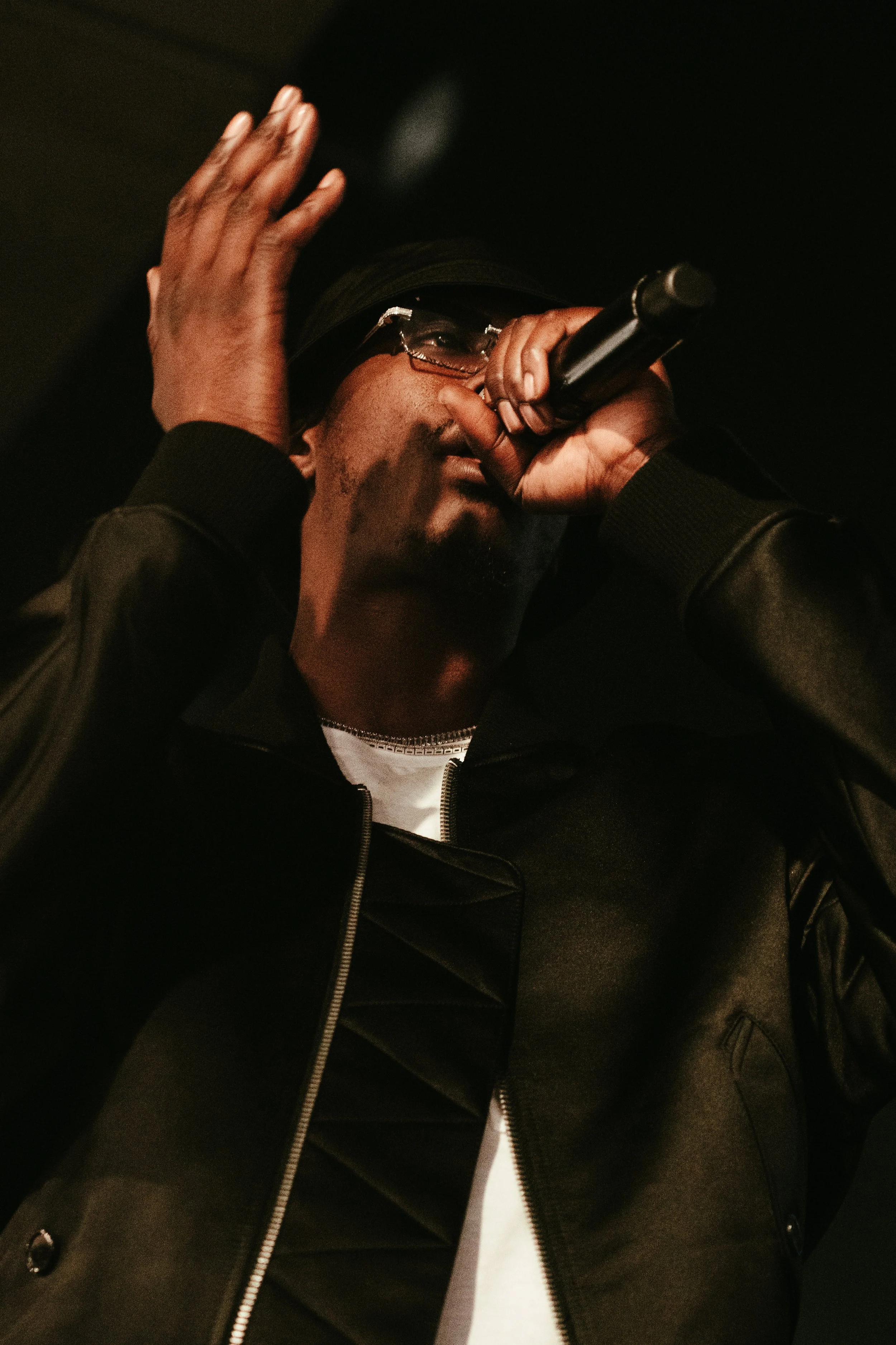 Un homme noir portant des lunettes, une casquette et un blouson noir, en train de chanter ou de parler dans un microphone.