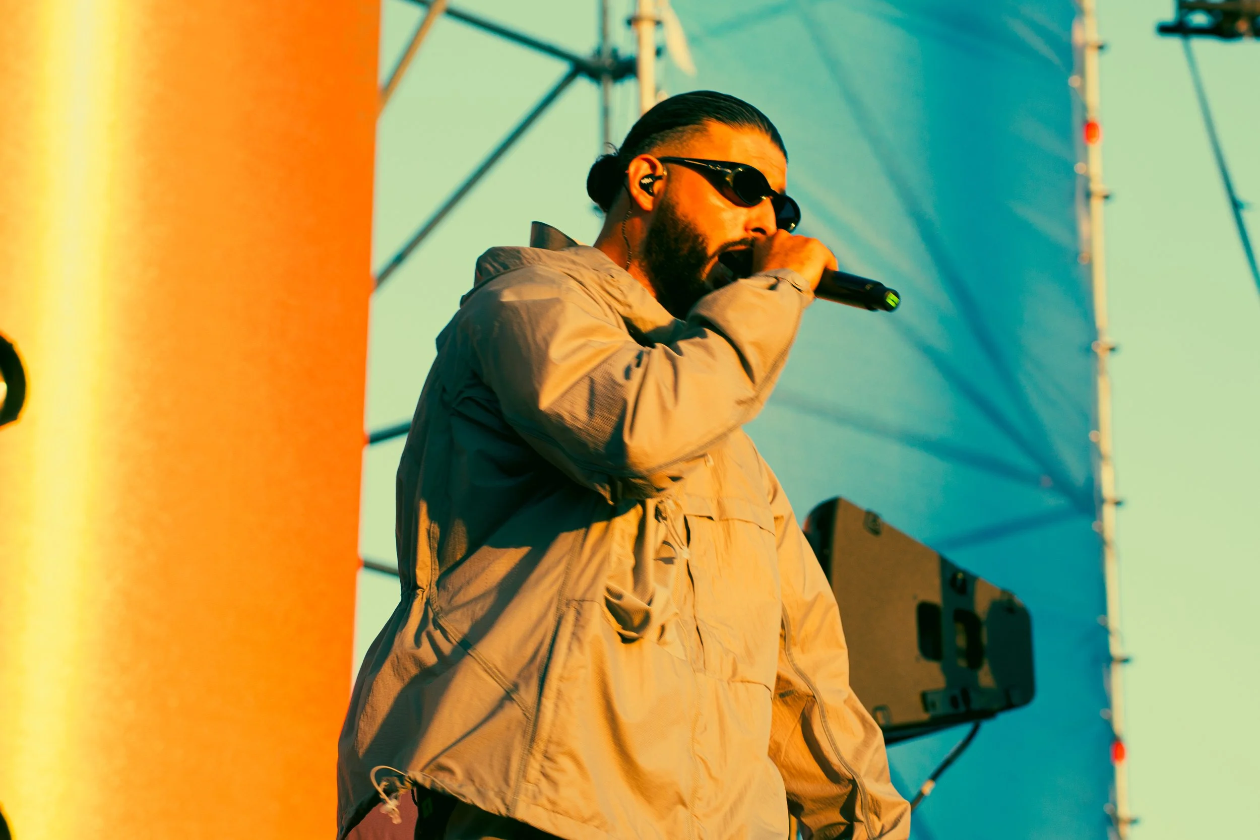 Un homme portant des lunettes de soleil, une veste beige, qui chante ou parle dans un micro lors d'un concert ou d'un événement en plein air.