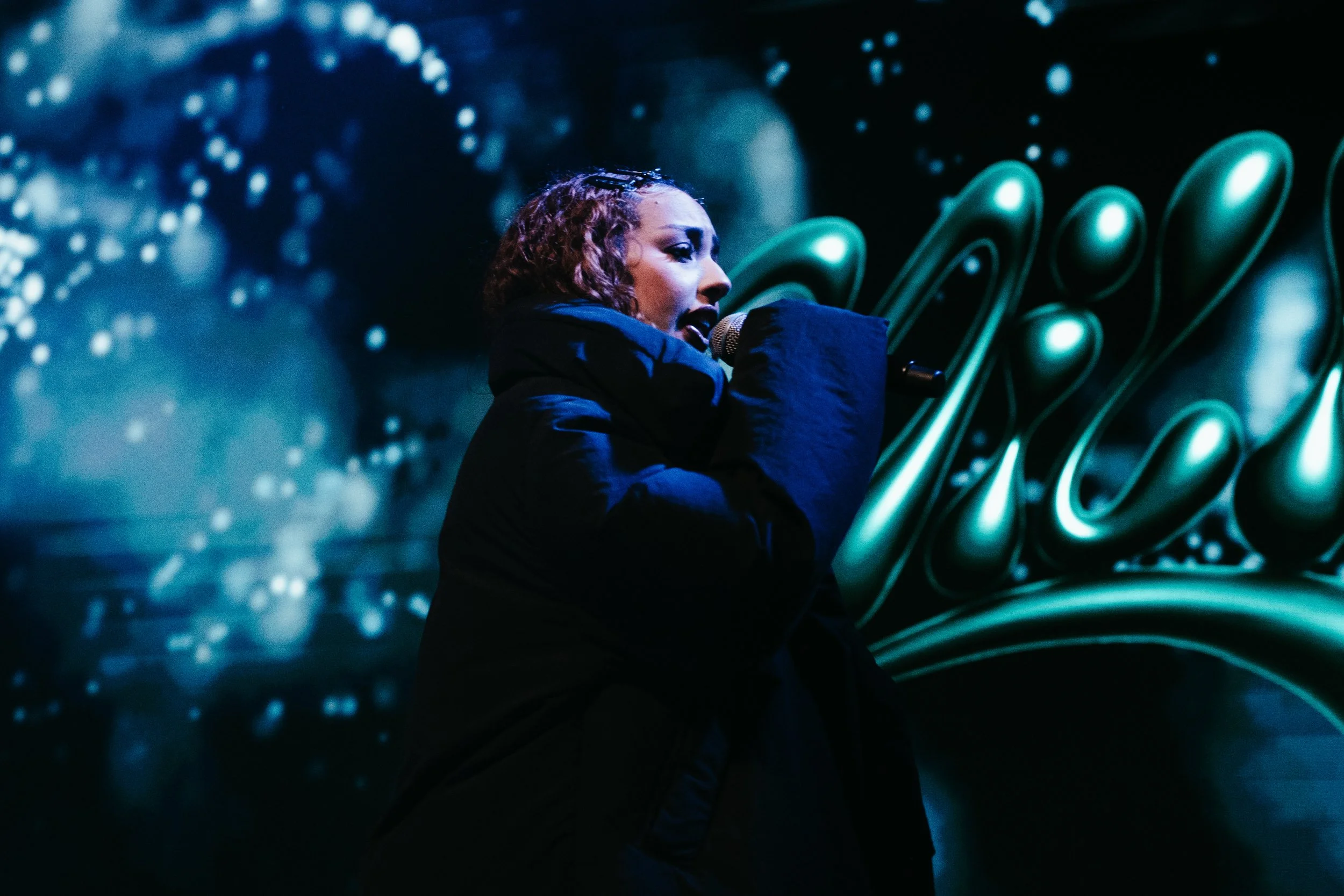 Une femme chante dans un micro, vêtue d'un vêtement noir, avec un fond artistique abstrait en couleurs bleu et vert.