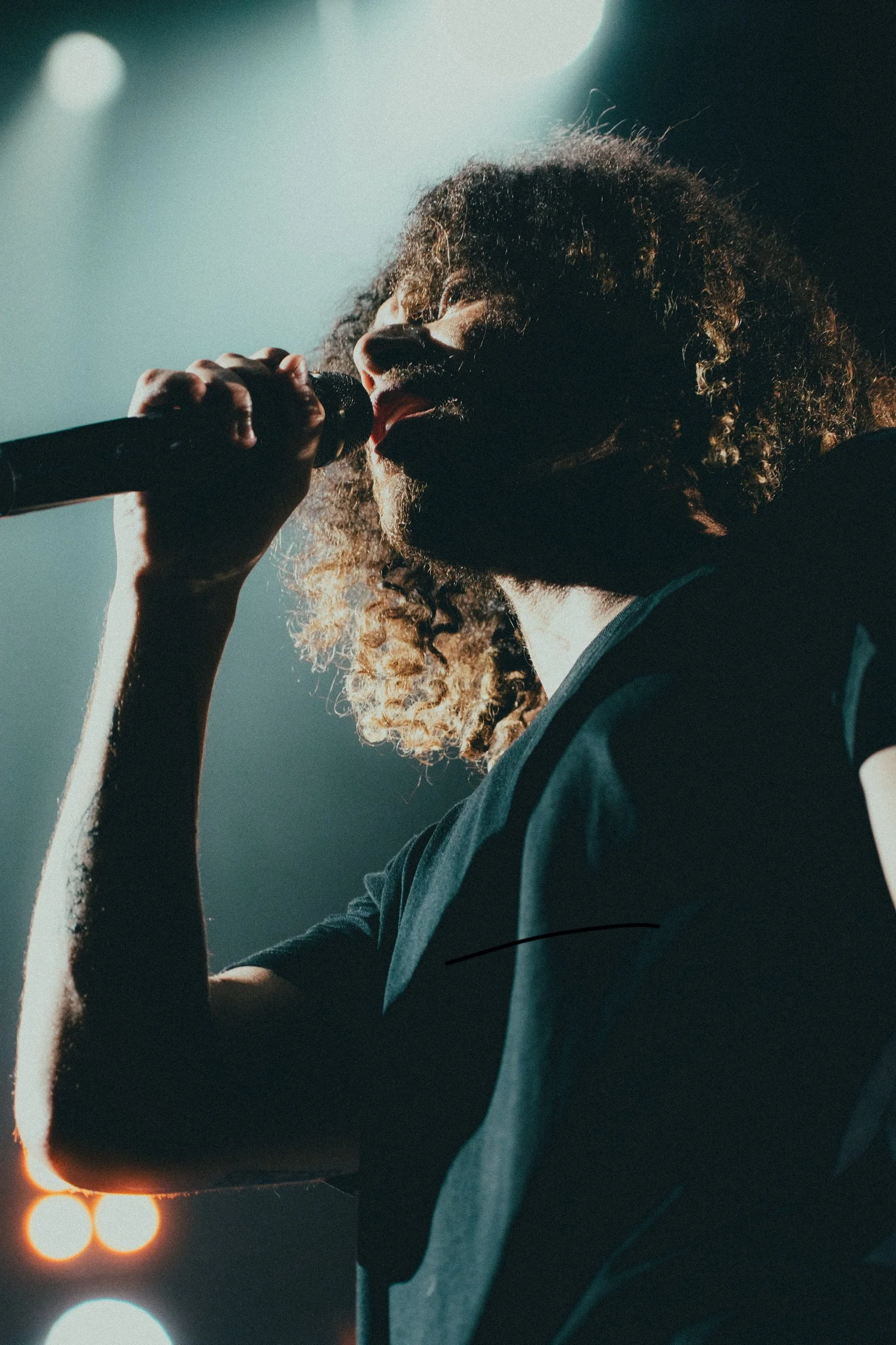 Un chanteur avec des cheveux crépus bruns, portant un t-shirt noir, tient un micro et chante sur scène, éclairé par des lumières de scène.