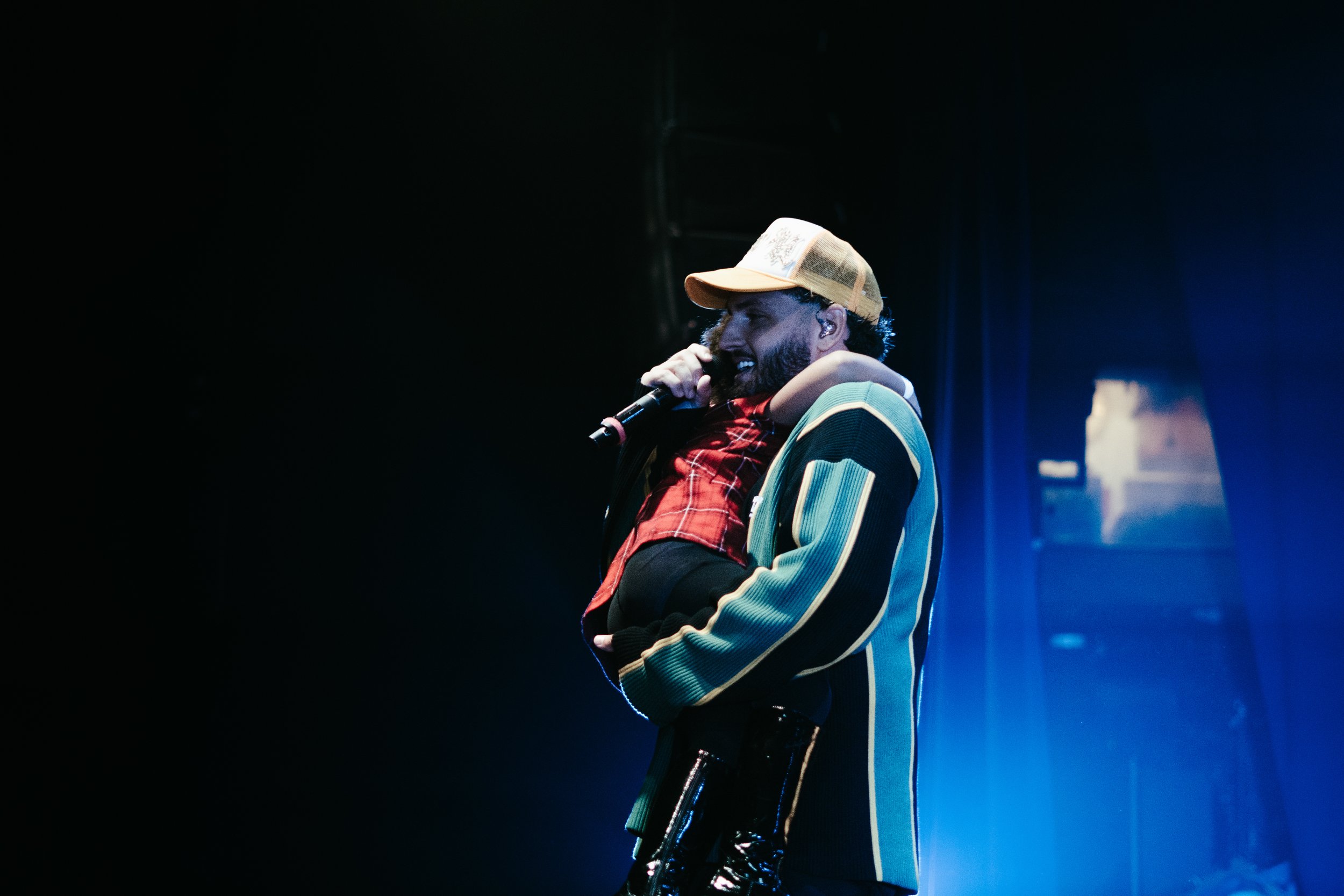 Un homme avec une barbe porte un jeune enfant sur ses épaules, tenant un microphone et chantant ou parlant sur scène. Il porte une casquette et un blouson à rayures, tandis que l'enfant porte une veste rouge à carreaux.