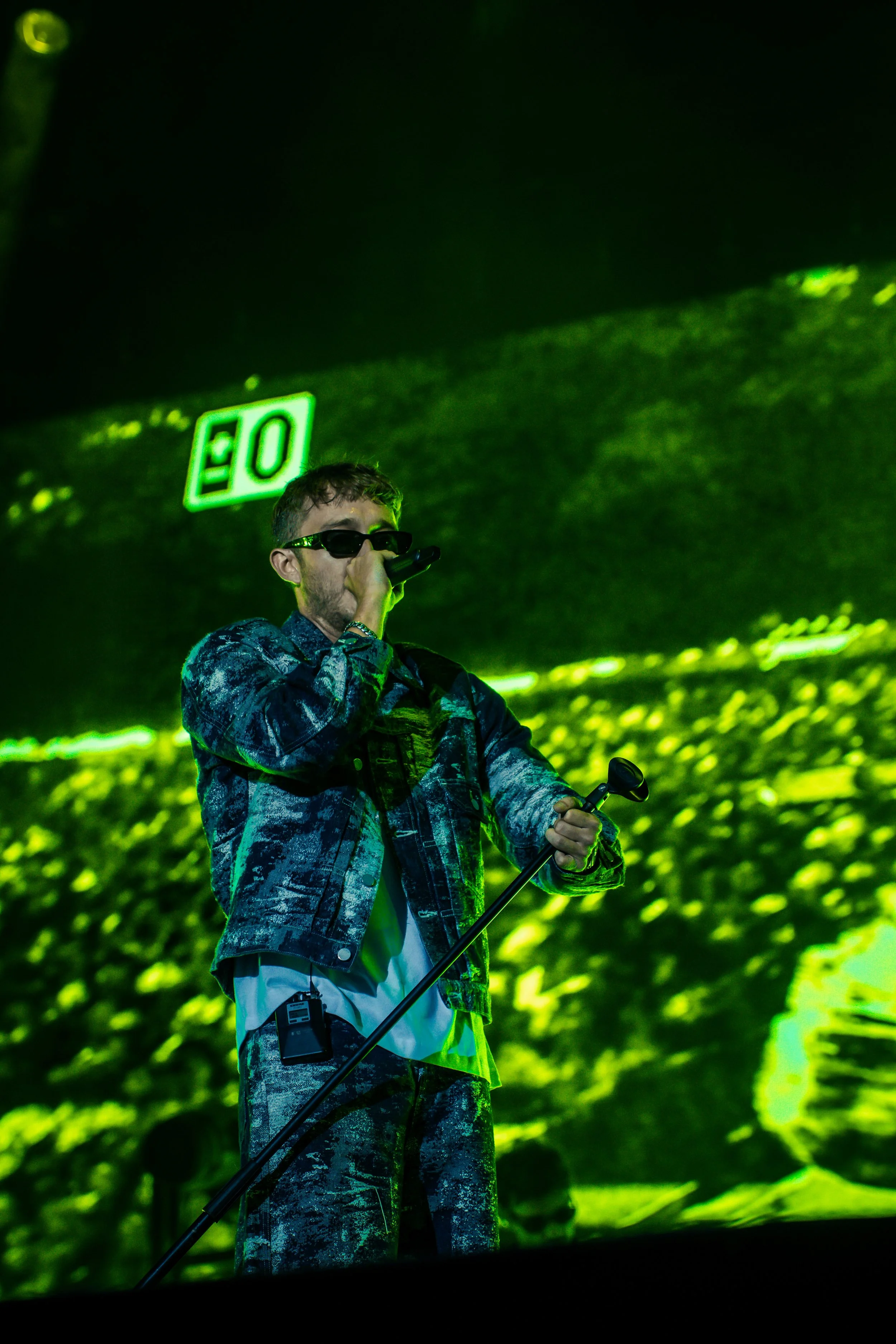 Un jeune homme portant des lunettes noires, une veste en denim et un pantalon camouflage, tient un microphone et un bâton de microphone lors d'une performance sur scène avec un fond lumineux vert.