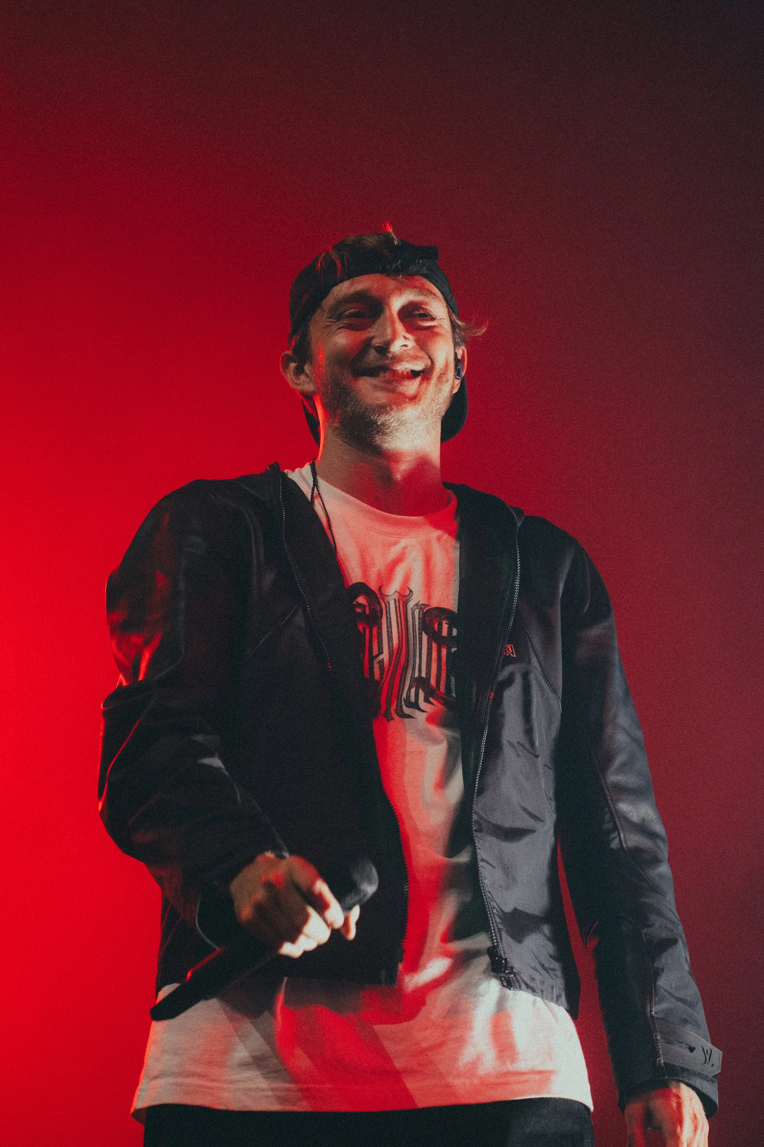 Homme souriant avec une casquette à l'envers, vêtement noir, joue avec un micro, fond rouge.