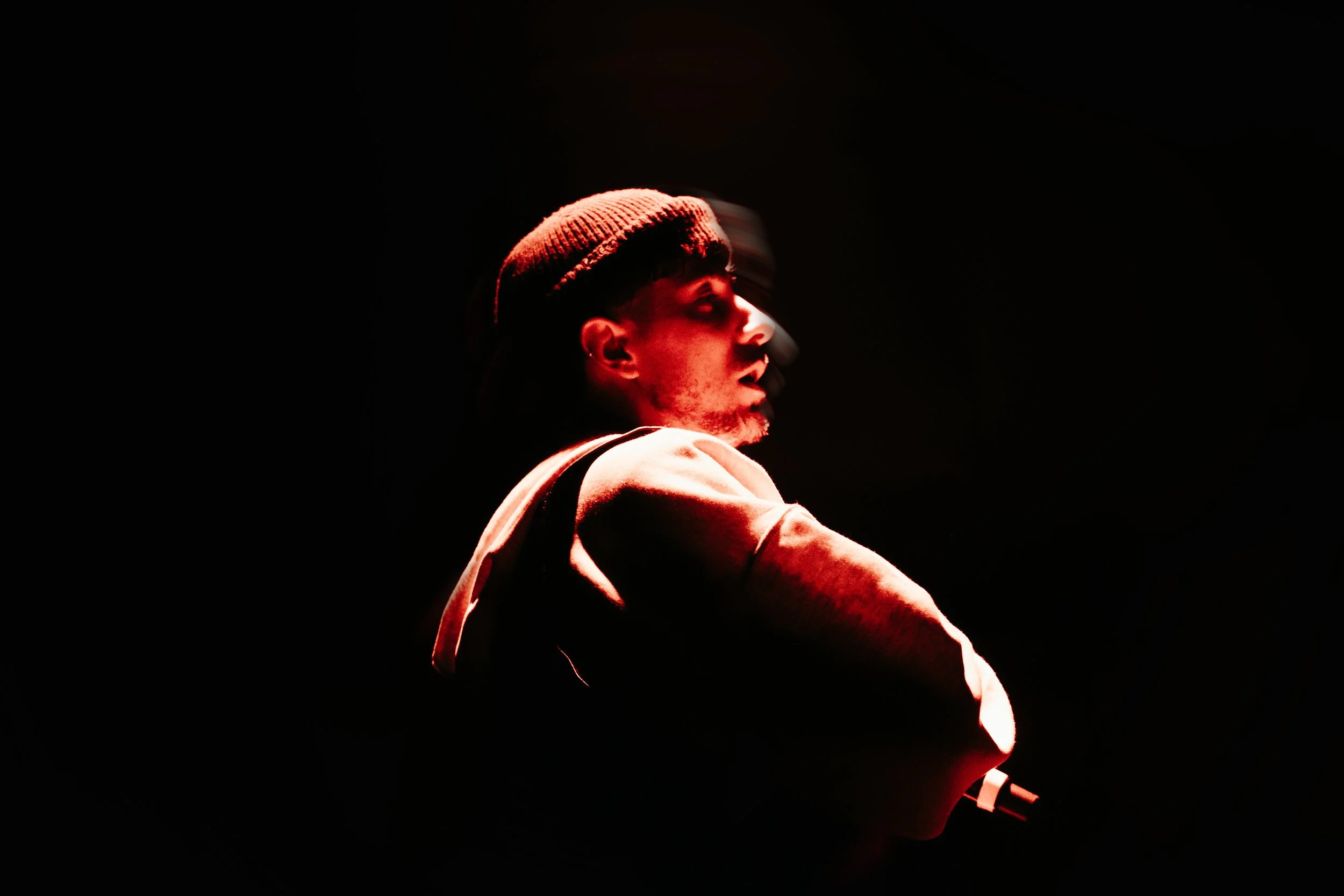 Portrait d'un homme portant une casquette et une veste, éclairé par une lumière rouge sur un fond noir.