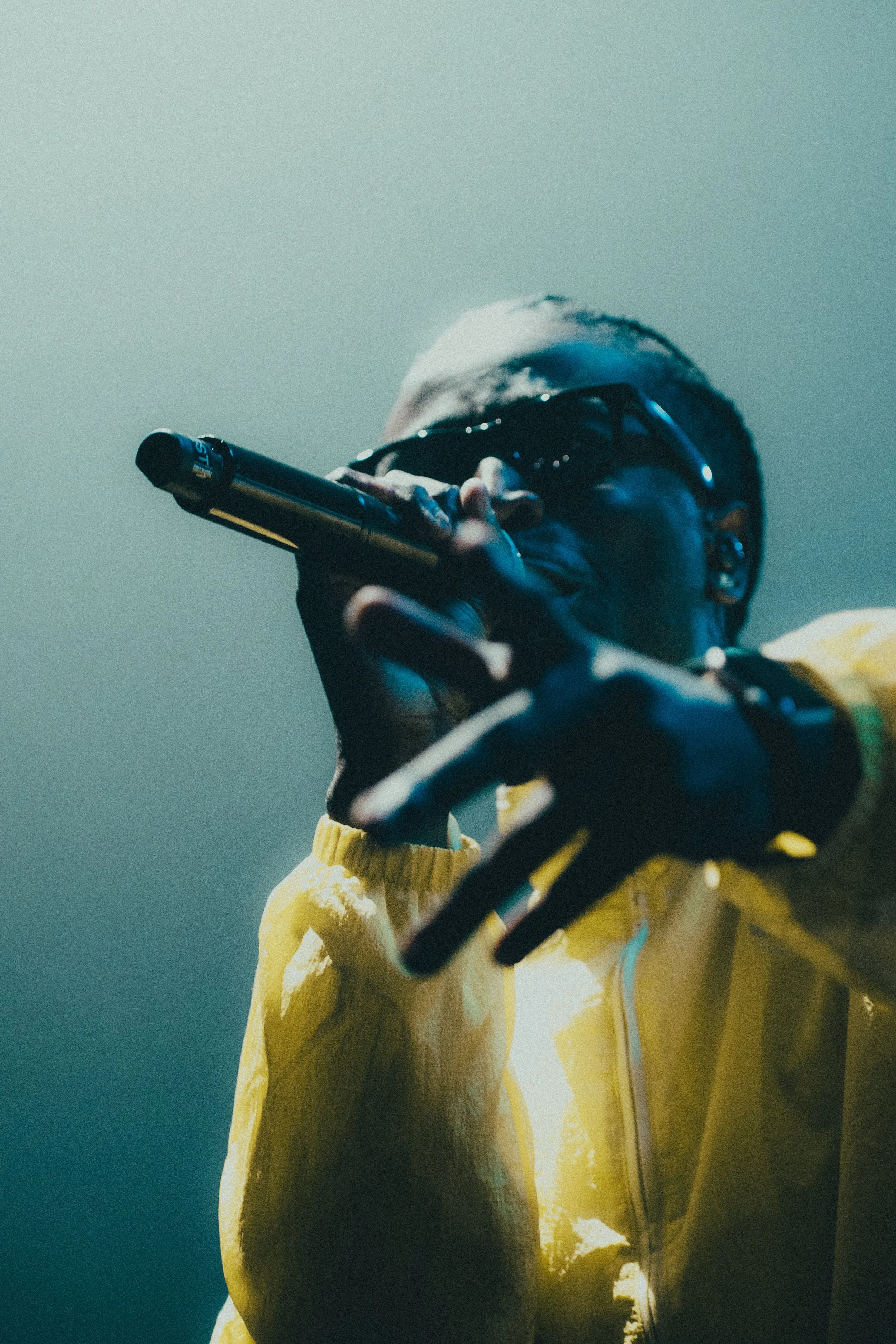 Un artiste de hip-hop portant des lunettes de soleil noires, un sweat jaune, et tenant un microphone noir, lors d'une performance sur scène.