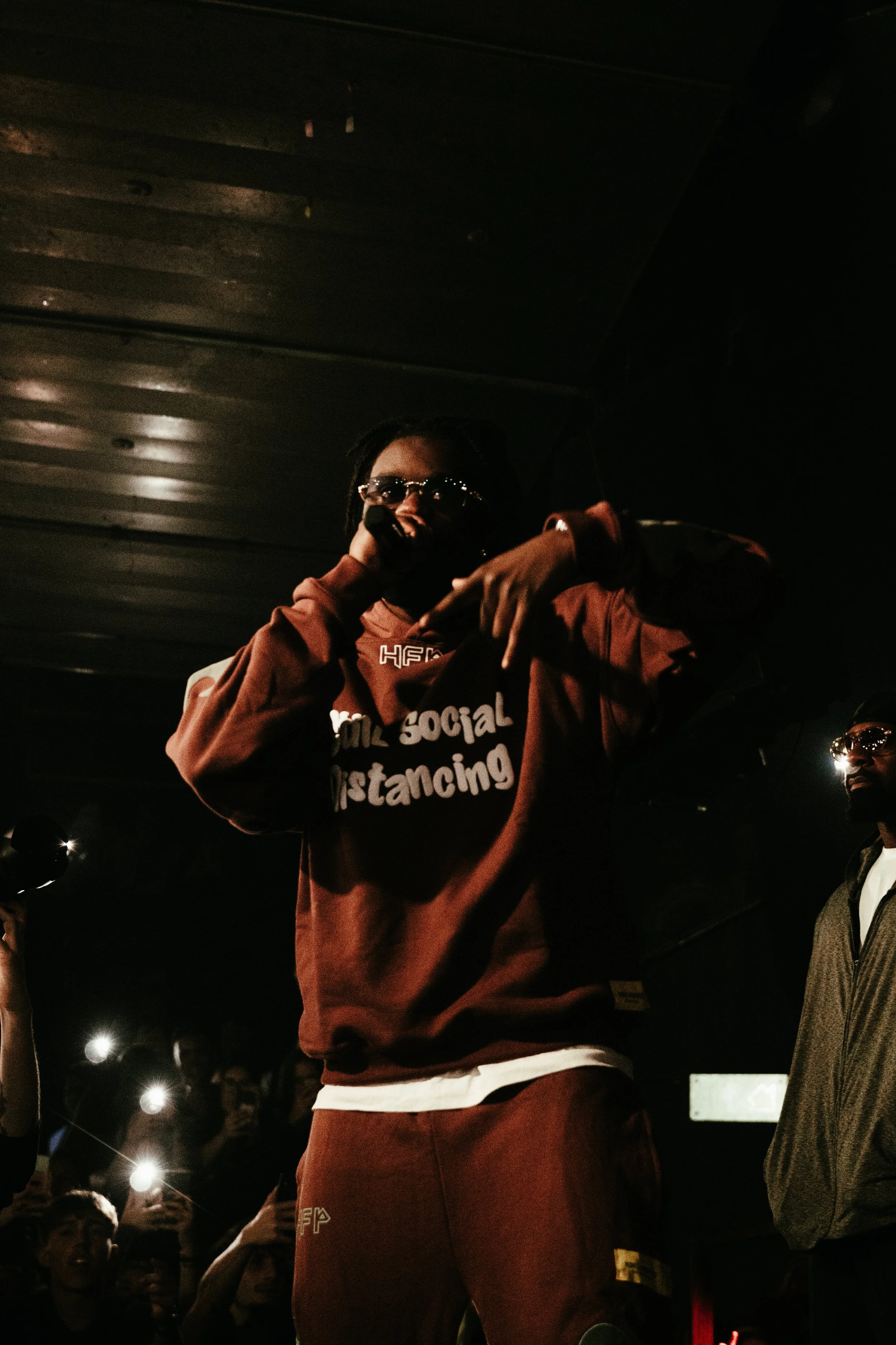 Un rappeur en hoodie marron performe sur scène, entouré de fans prenant des photos avec leurs téléphones, dans un environnement sombre.